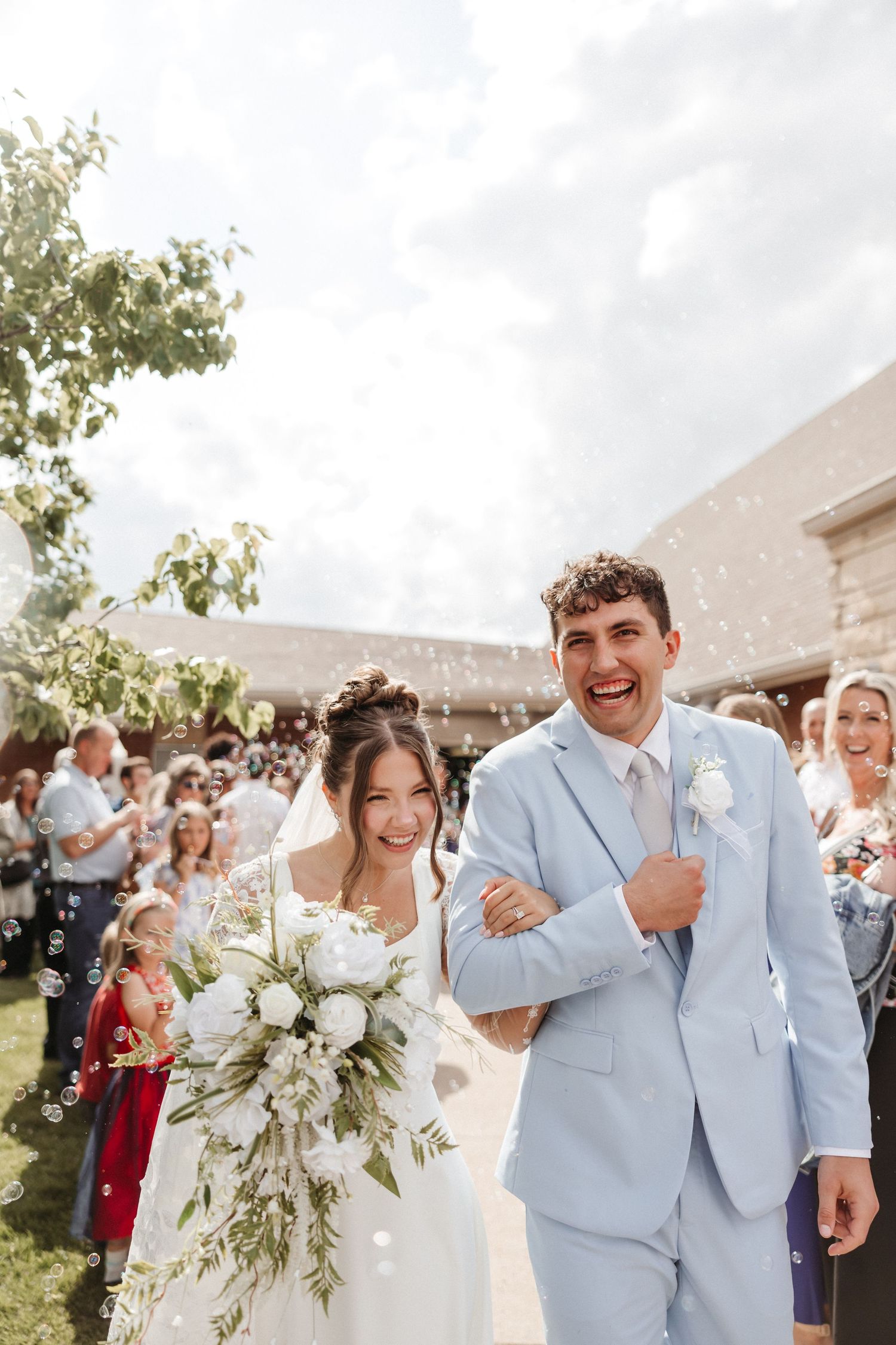 Joyful newlyweds walk through bubbles, bride holding white bouquet in wedding dress, groom in light blue suit.