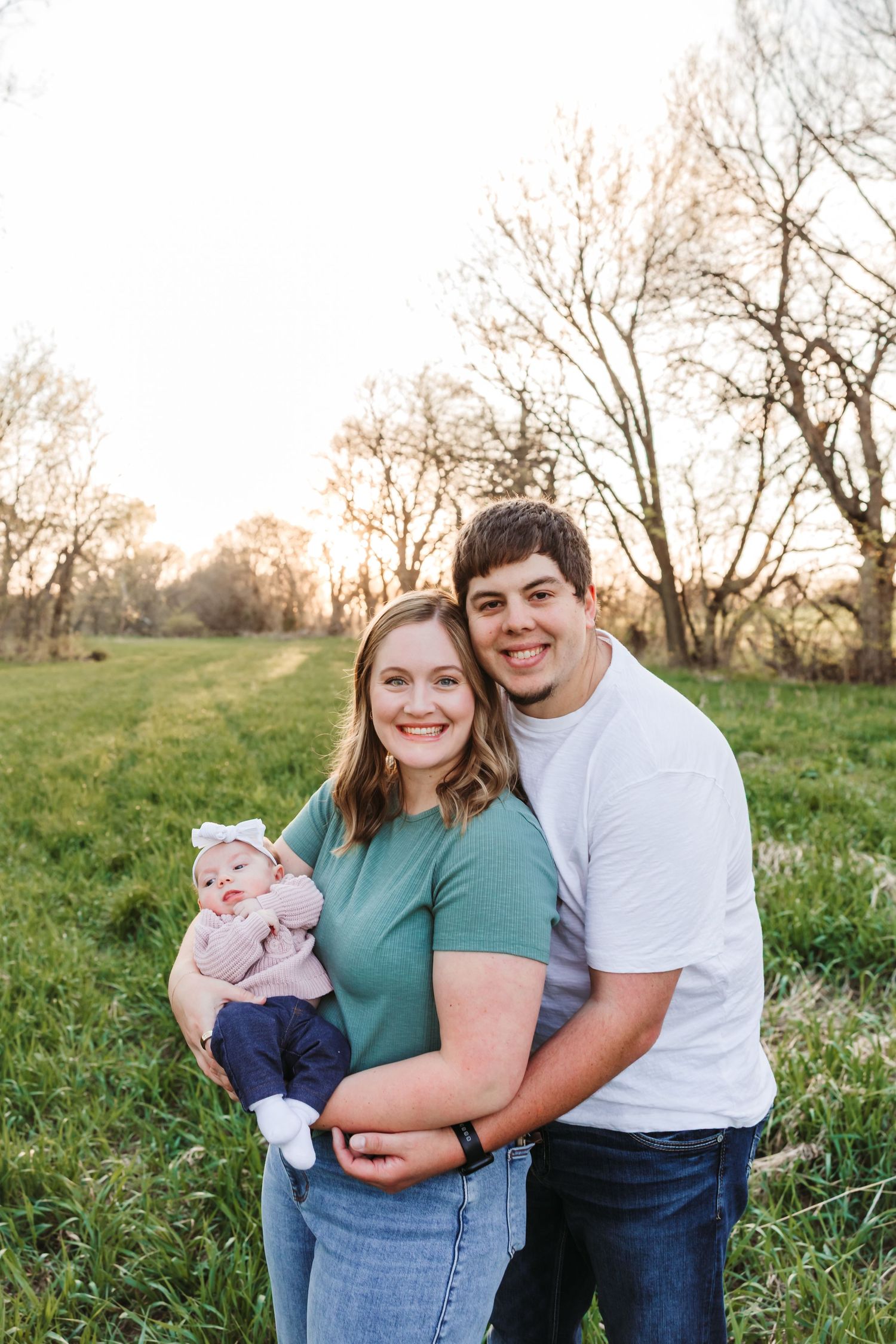 A young couple smiles outdoors holding their baby in a green field with bare trees at golden hour.