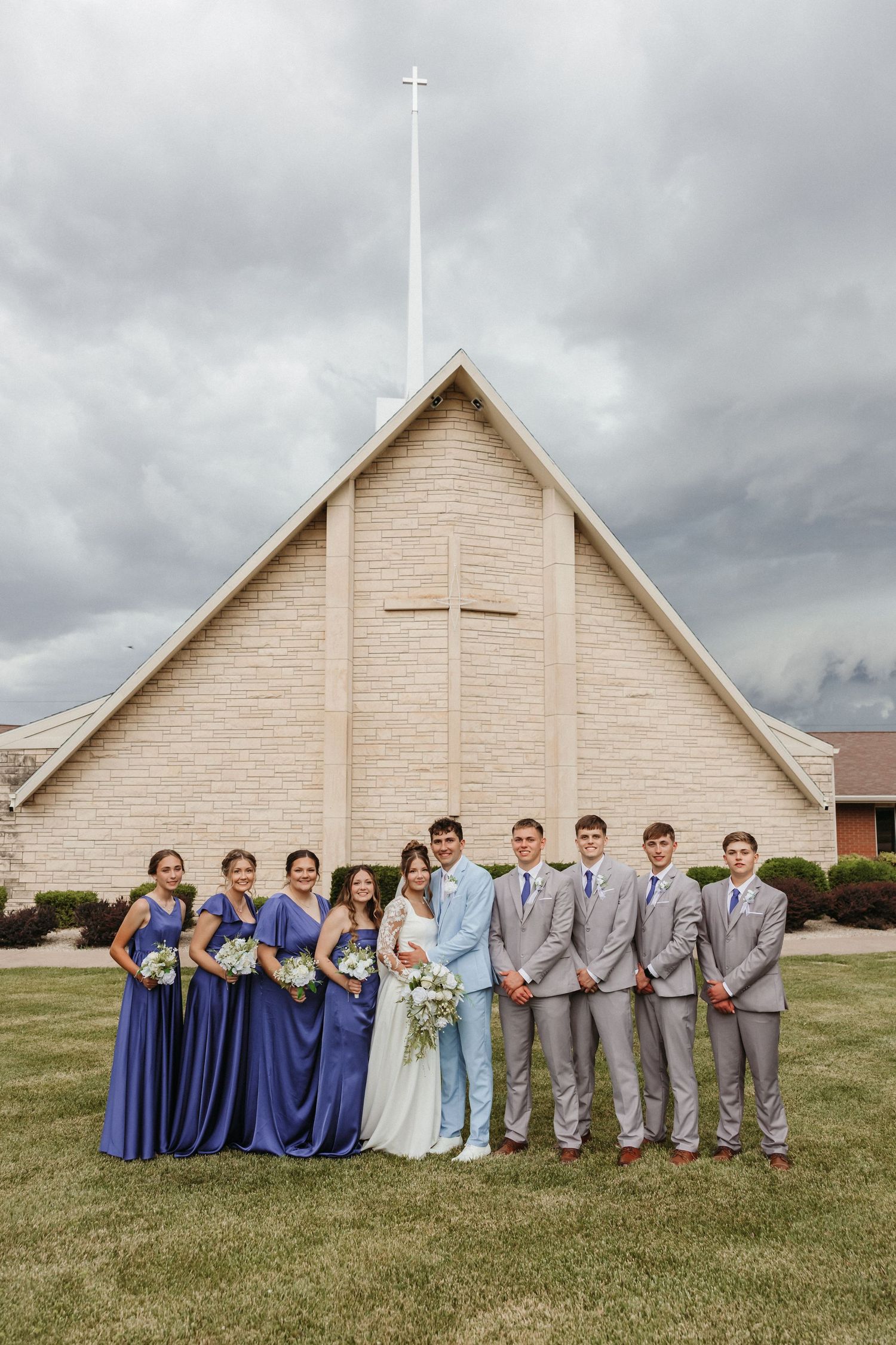 Wedding party poses before a church, bridesmaids in blue gowns and groomsmen in gray suits flanking the bride and groom.