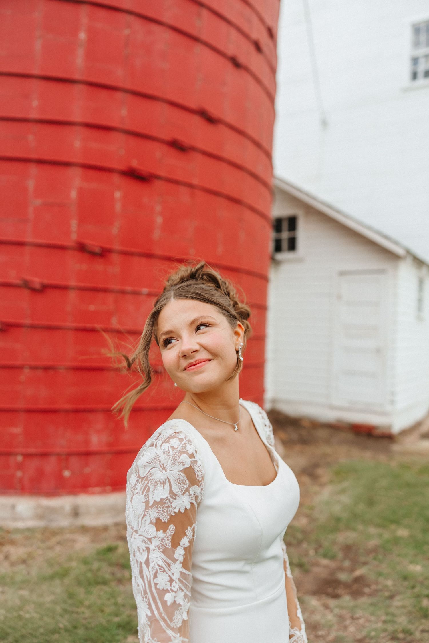 Smiling bride in a white lace long-sleeve wedding dress poses in front of a rustic red barn silo.
