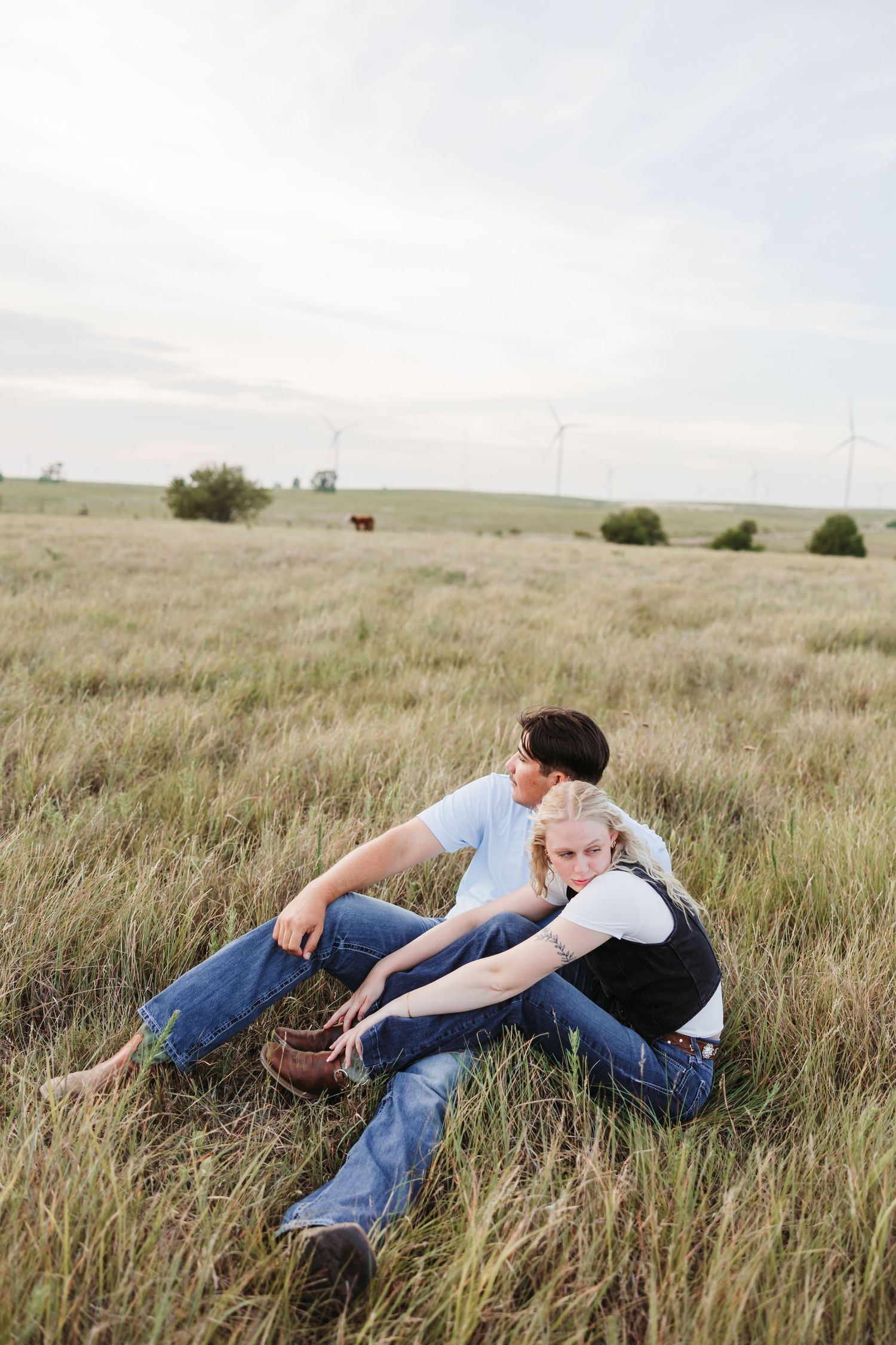 A couple relaxes in tall prairie grass with wind turbines visible in the distance.