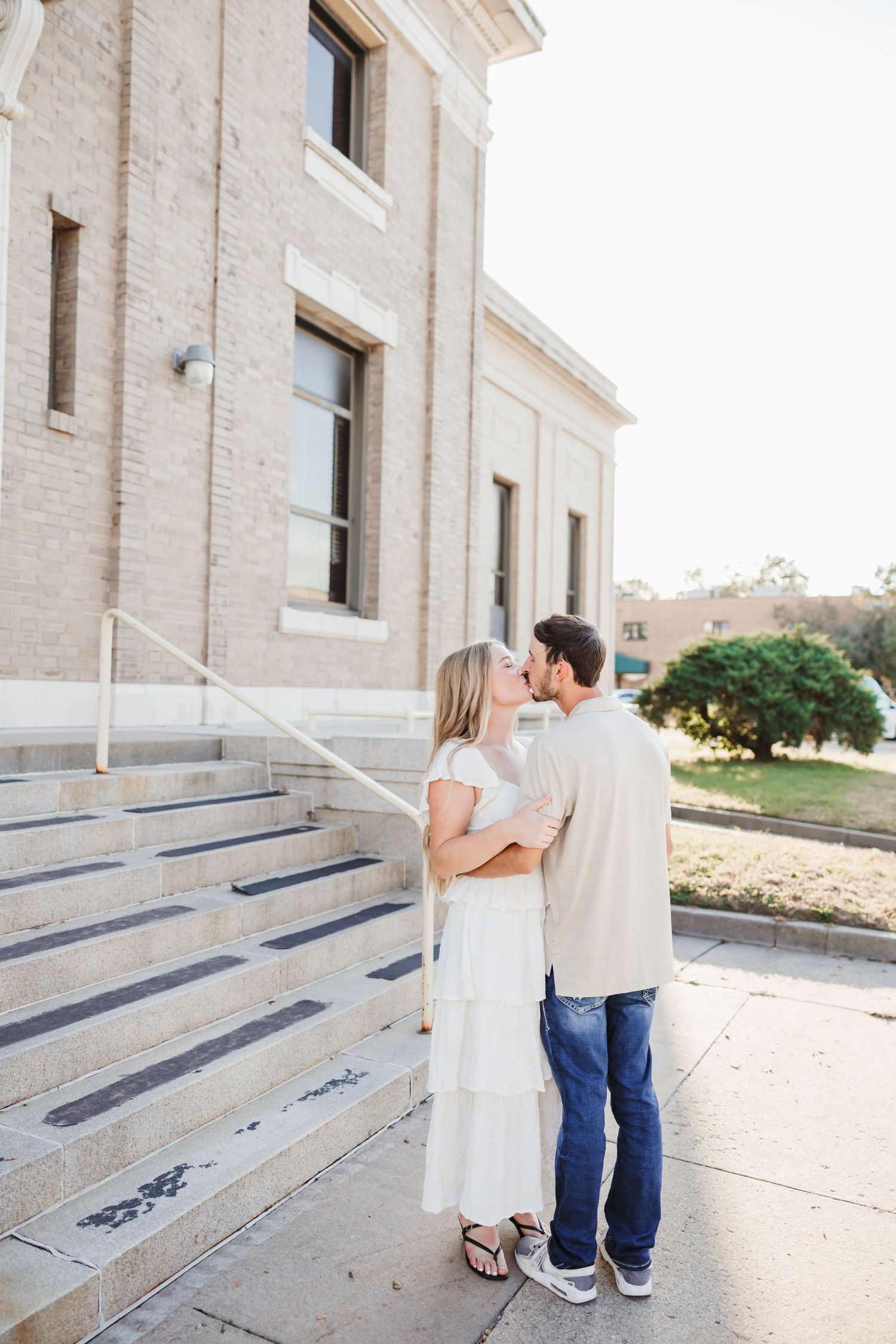 A couple embraces on stone steps outside a classic brick building.