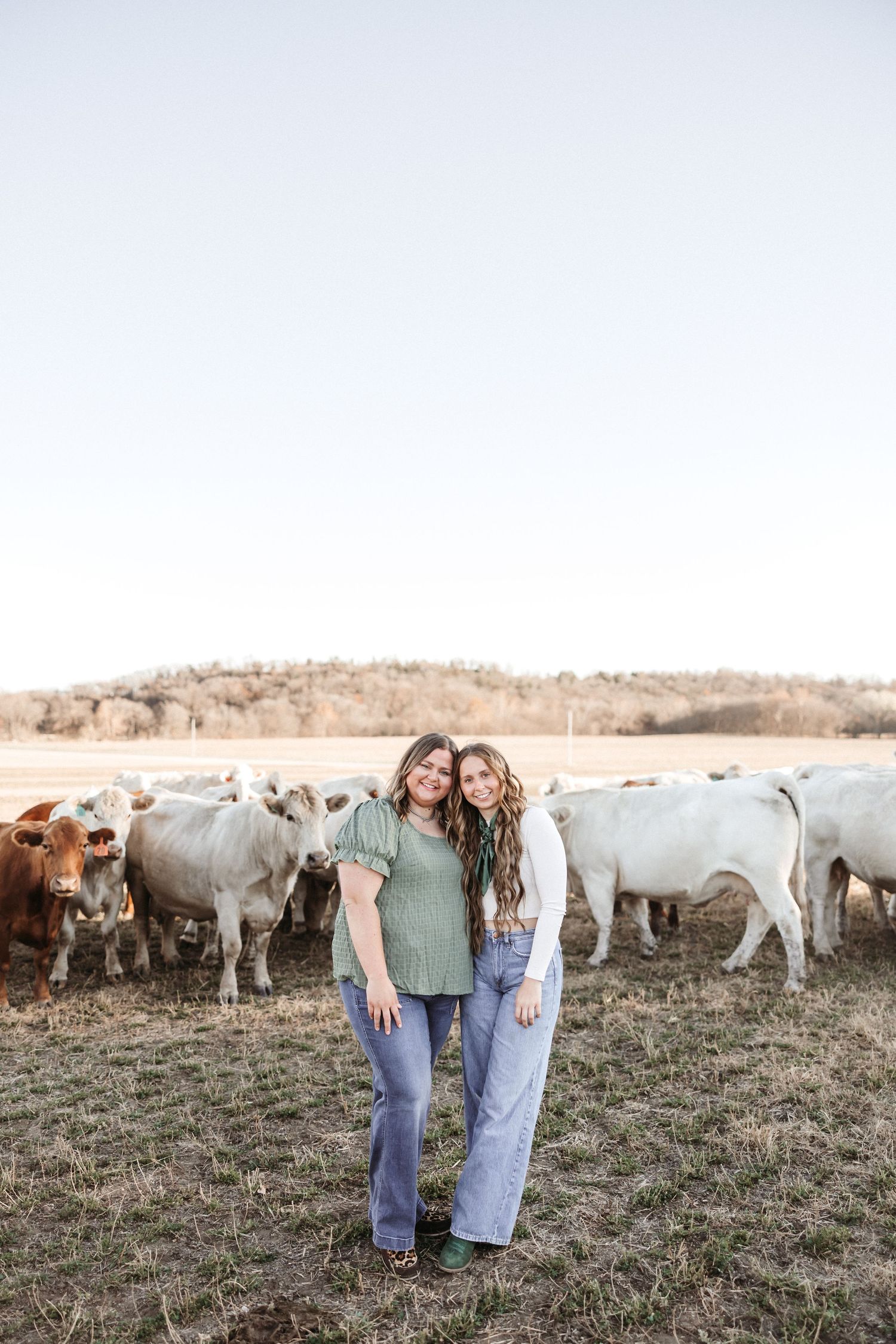 Two sisters pose together in styled attire with a herd of cattle visible behind them.
