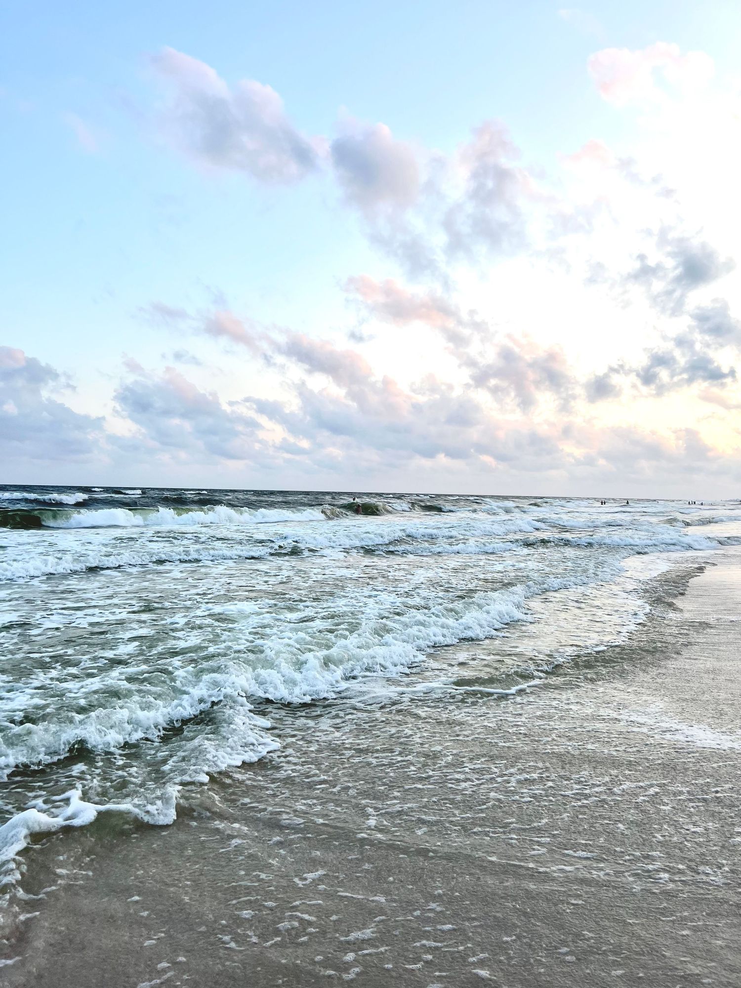 Gentle ocean waves washing onto a sandy beach under a pastel blue sky with soft pink clouds at dusk.