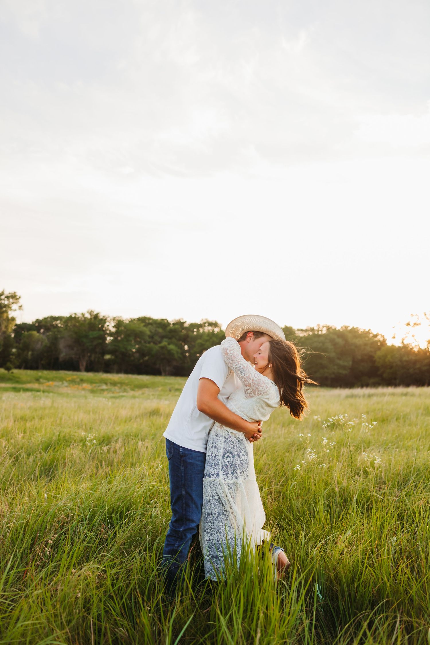 Couple embracing and kissing in a lush green field at sunset, woman in white lace dress and man in cowboy hat.