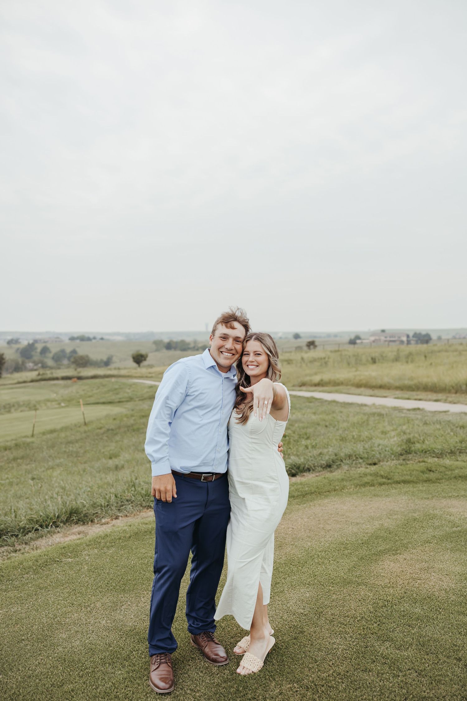A happy couple poses together on a scenic golf course overlooking rolling hills after becoming engaged.