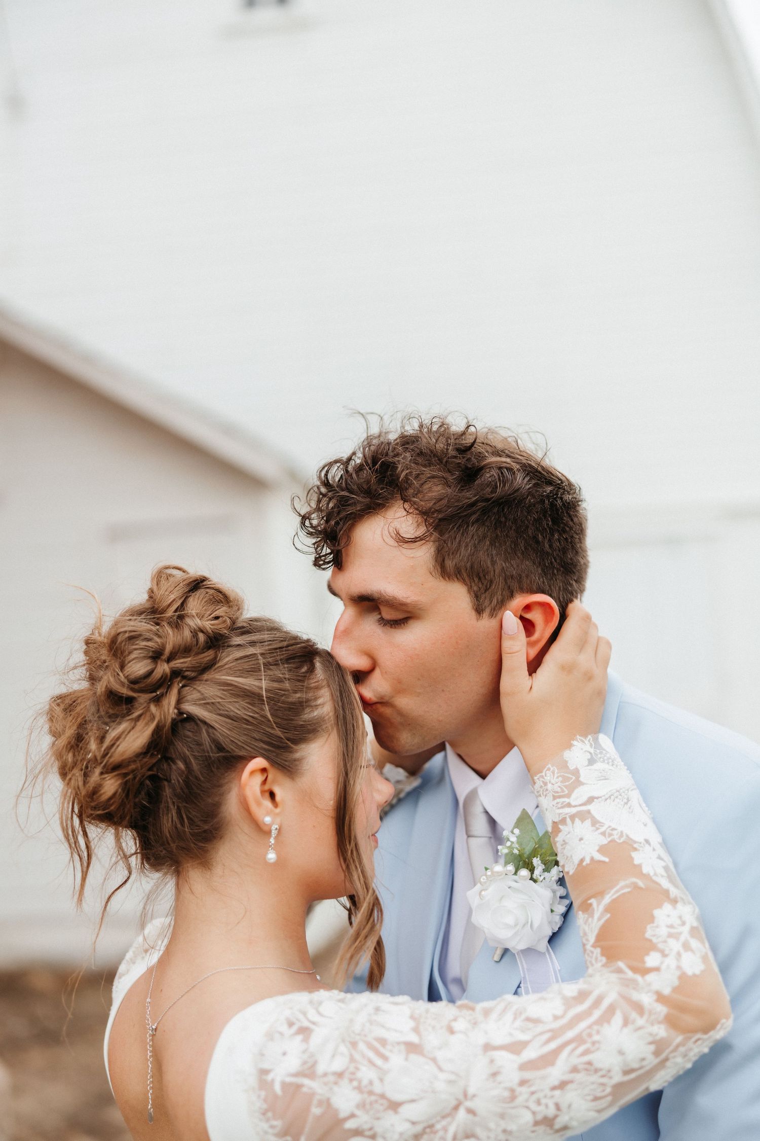 A bride and groom share a romantic kiss outdoors, with the groom in a light blue suit and boutonniere.