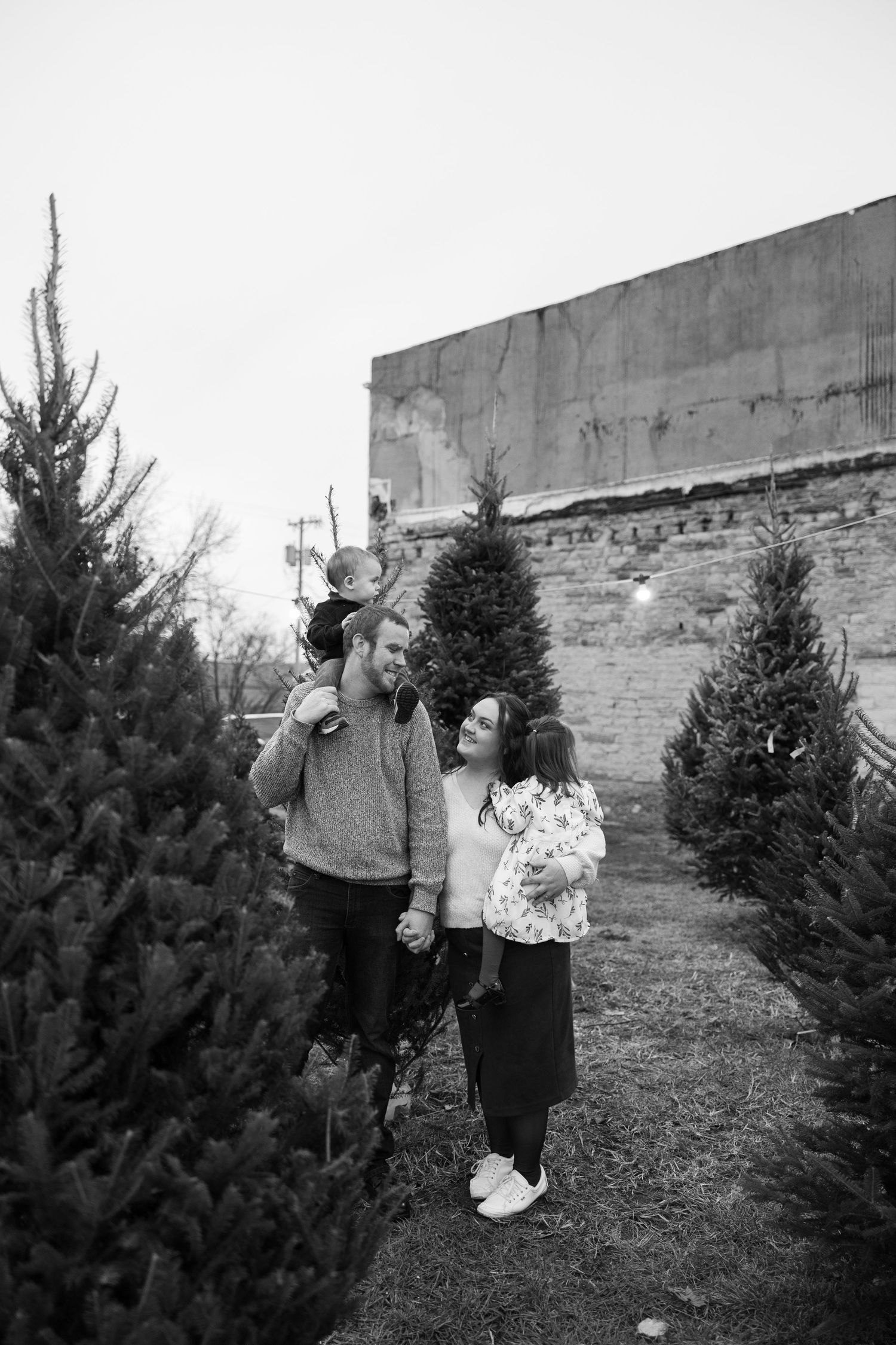 People browsing Christmas trees at an outdoor tree lot with brick wall background.