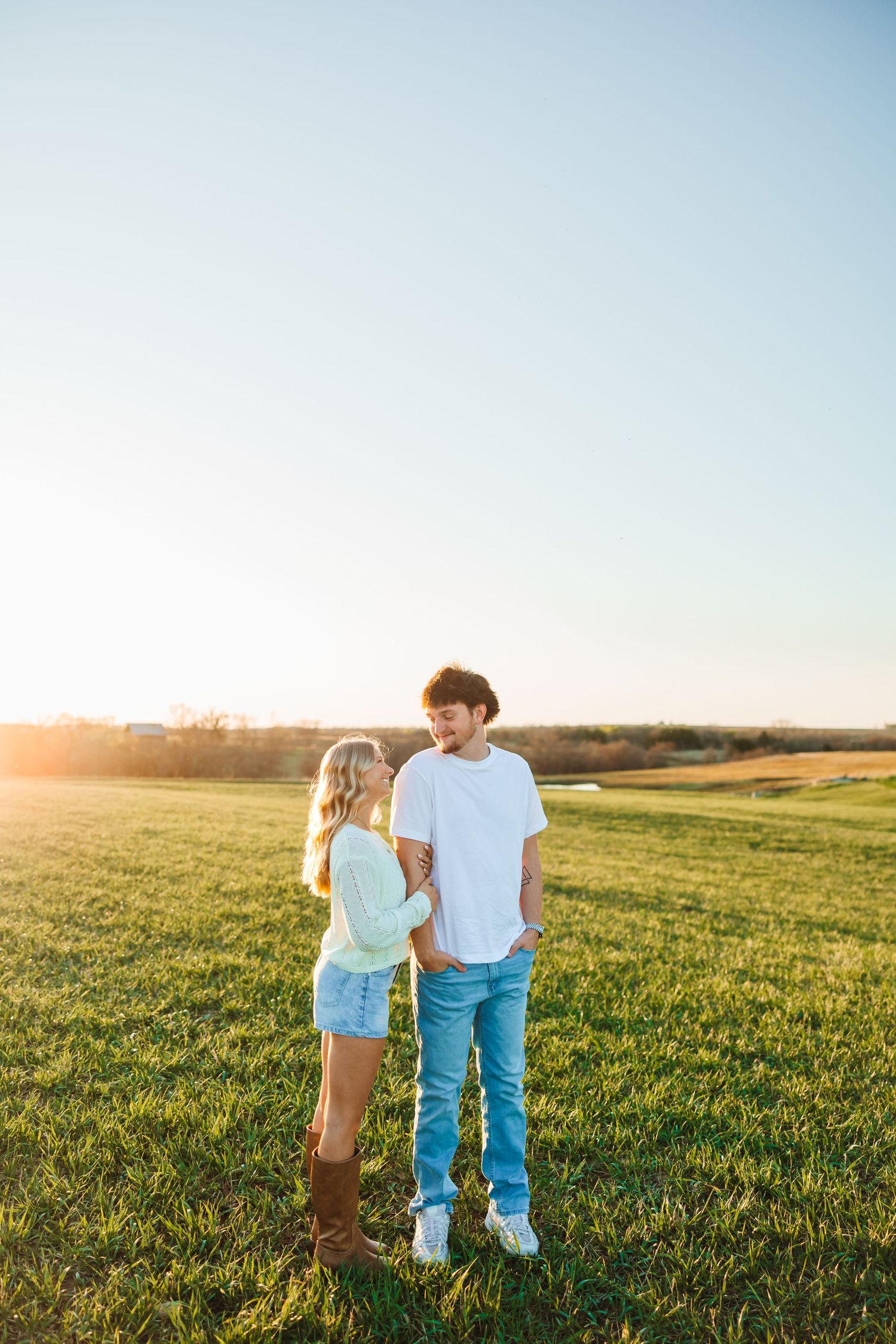 A couple stands in a sunlit green field at golden hour, smiling at each other in casual summer outfits.