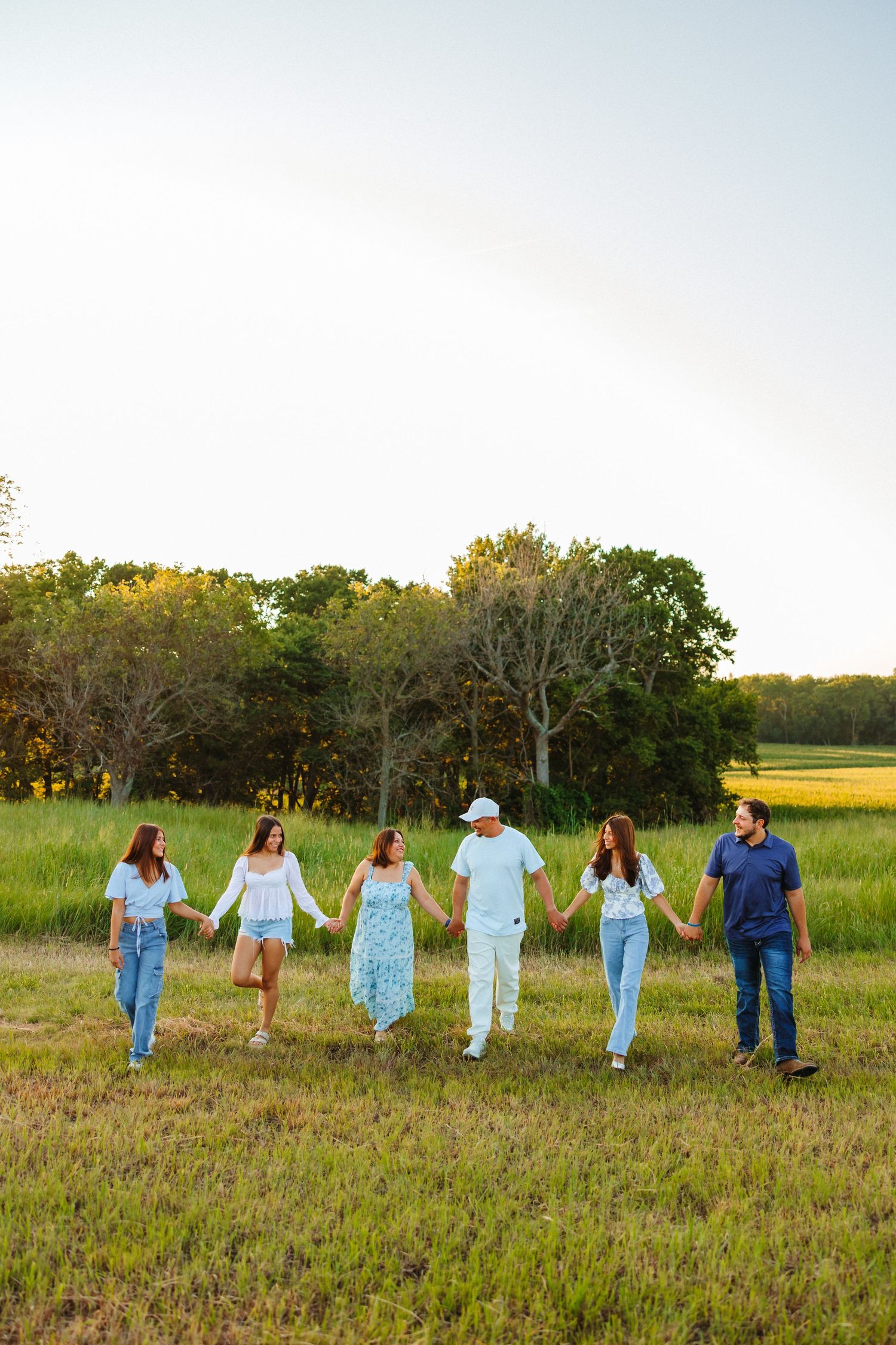 Six friends hold hands walking through a green field at golden hour with trees in the background.