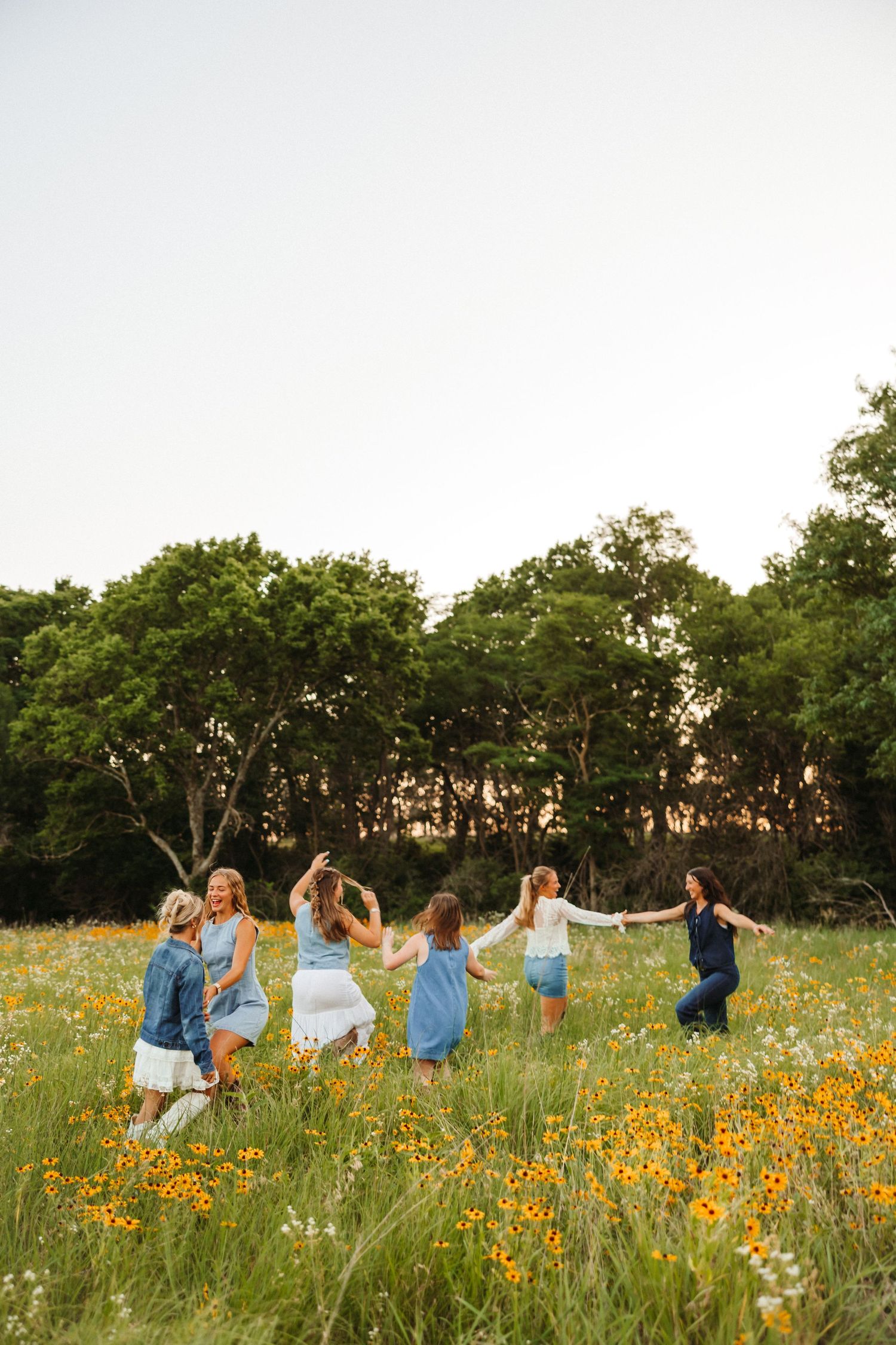Group of women in blue and white dresses dancing in a wildflower meadow with trees in the background.