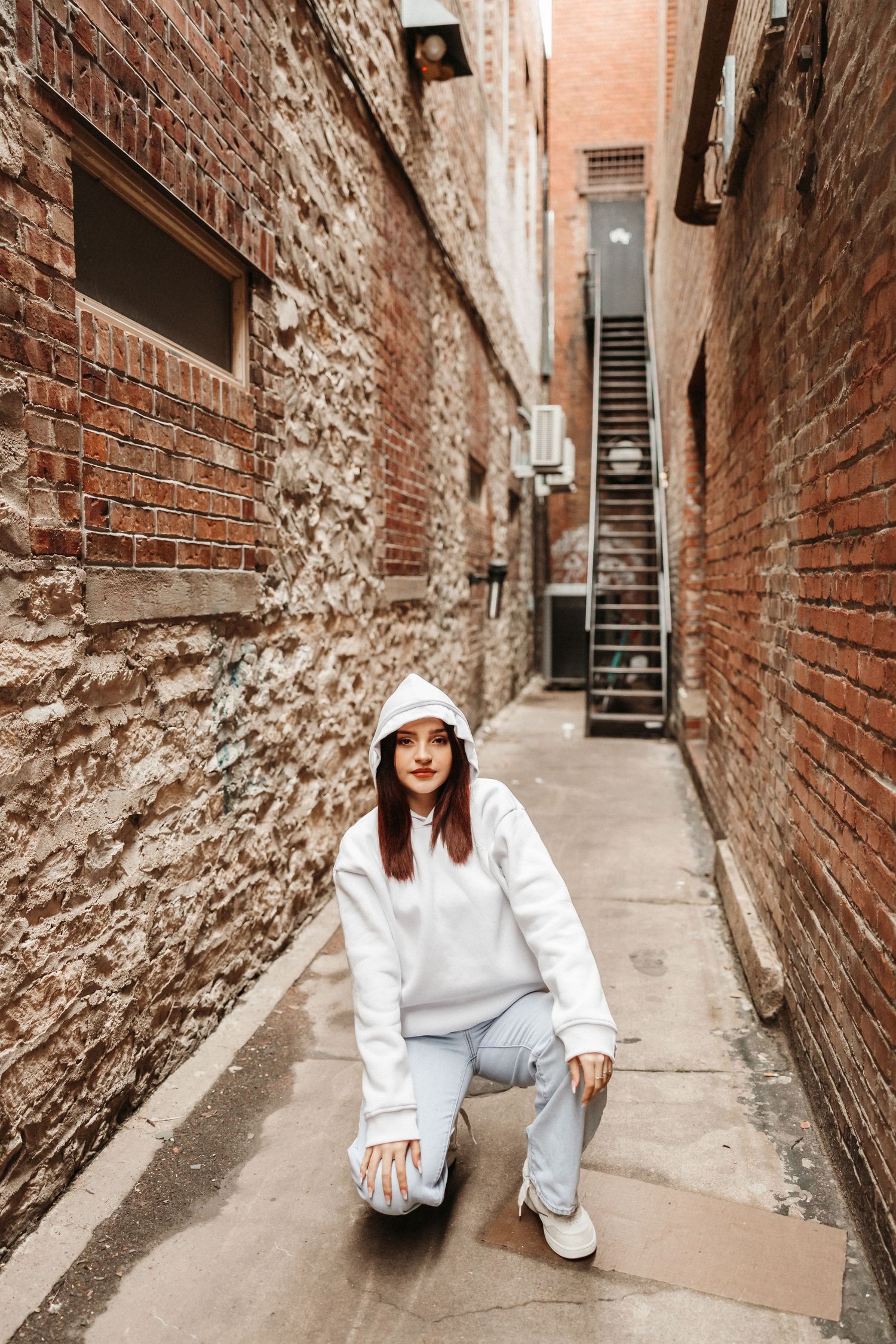 Young woman in white hoodie and light blue pants crouching in a narrow urban brick alley with stairs in background.