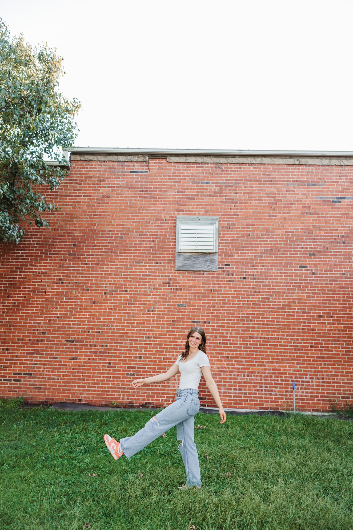 Young woman in white top and jeans posing playfully on grass in front of a red brick wall outdoors.