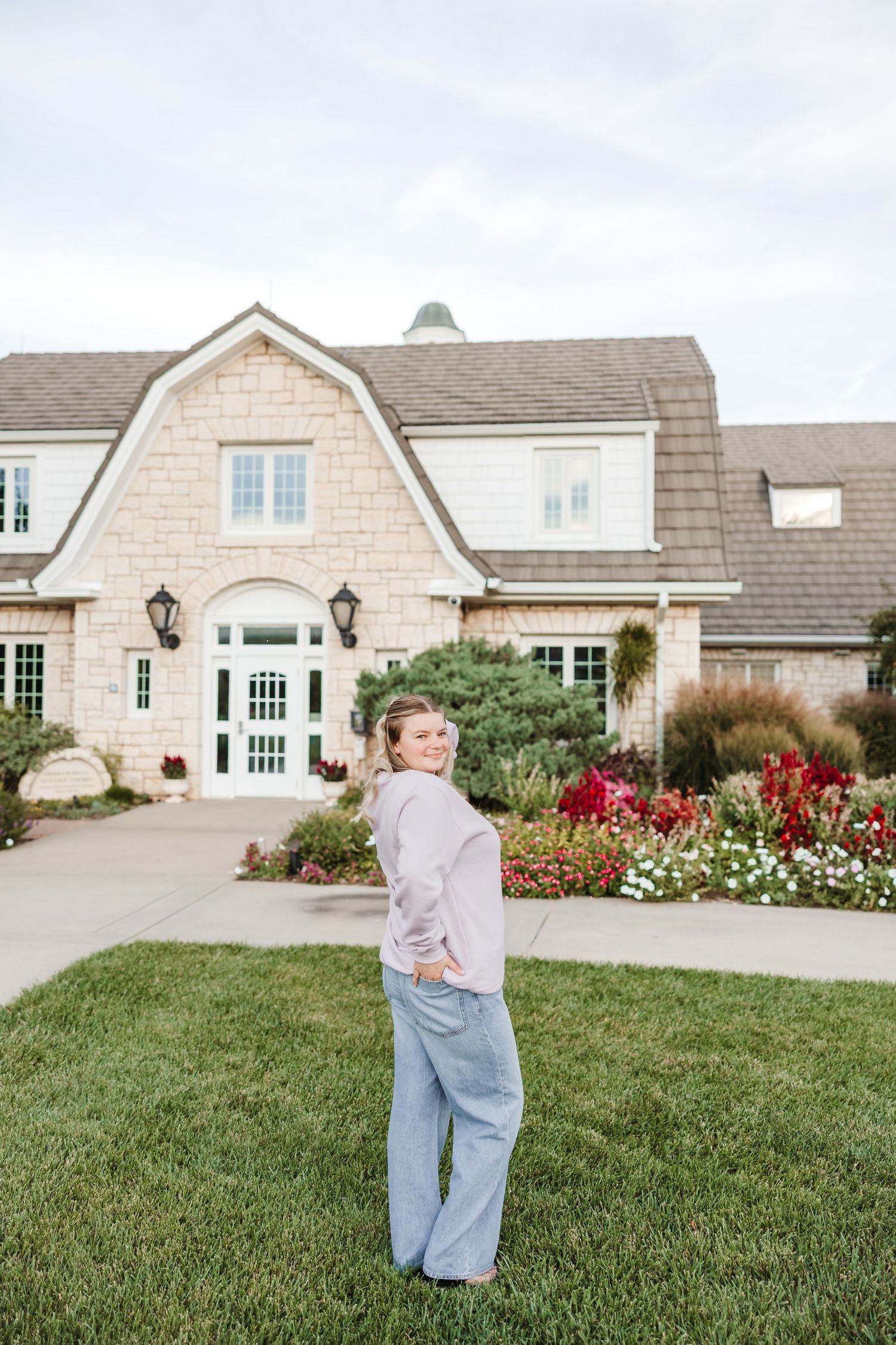 Woman in pink top and gray pants smiling in front of a stone house with colorful flower gardens.