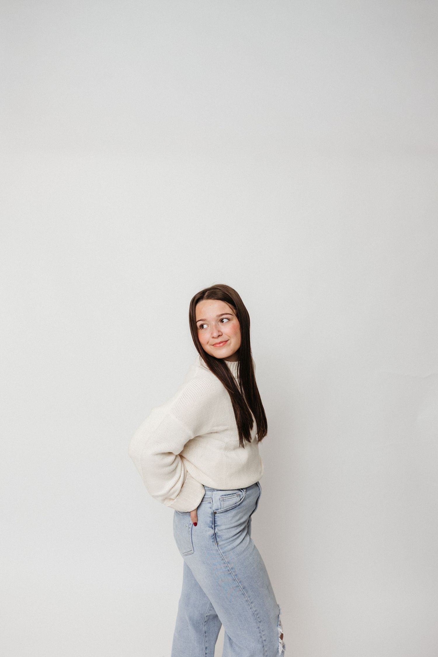 Stylish pose in cream sweater and light denim against minimalist white backdrop.