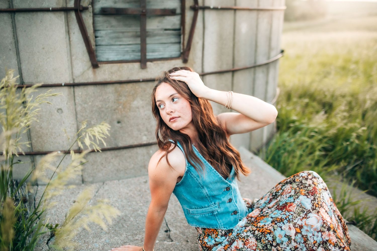 Woman in denim top and floral skirt poses gracefully by rustic building at sunset.