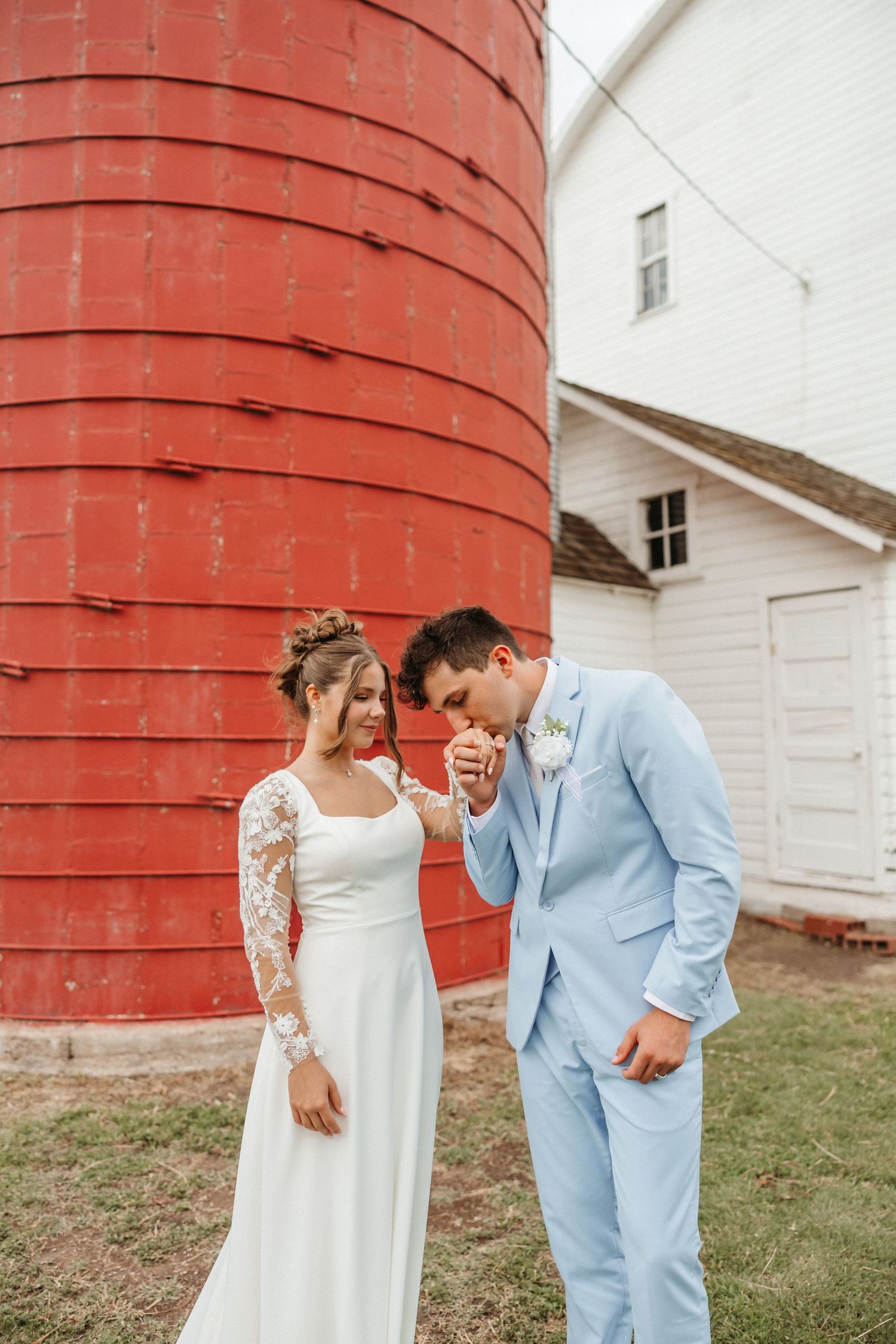 Bride and groom pose by a red silo, groom in light blue suit kissing bride's hand in her lace wedding dress.