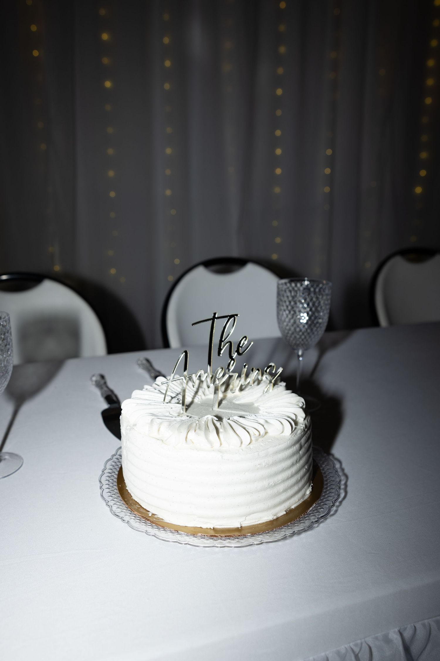 Elegant white wedding cake with silver topper on a white tablecloth at a reception venue.