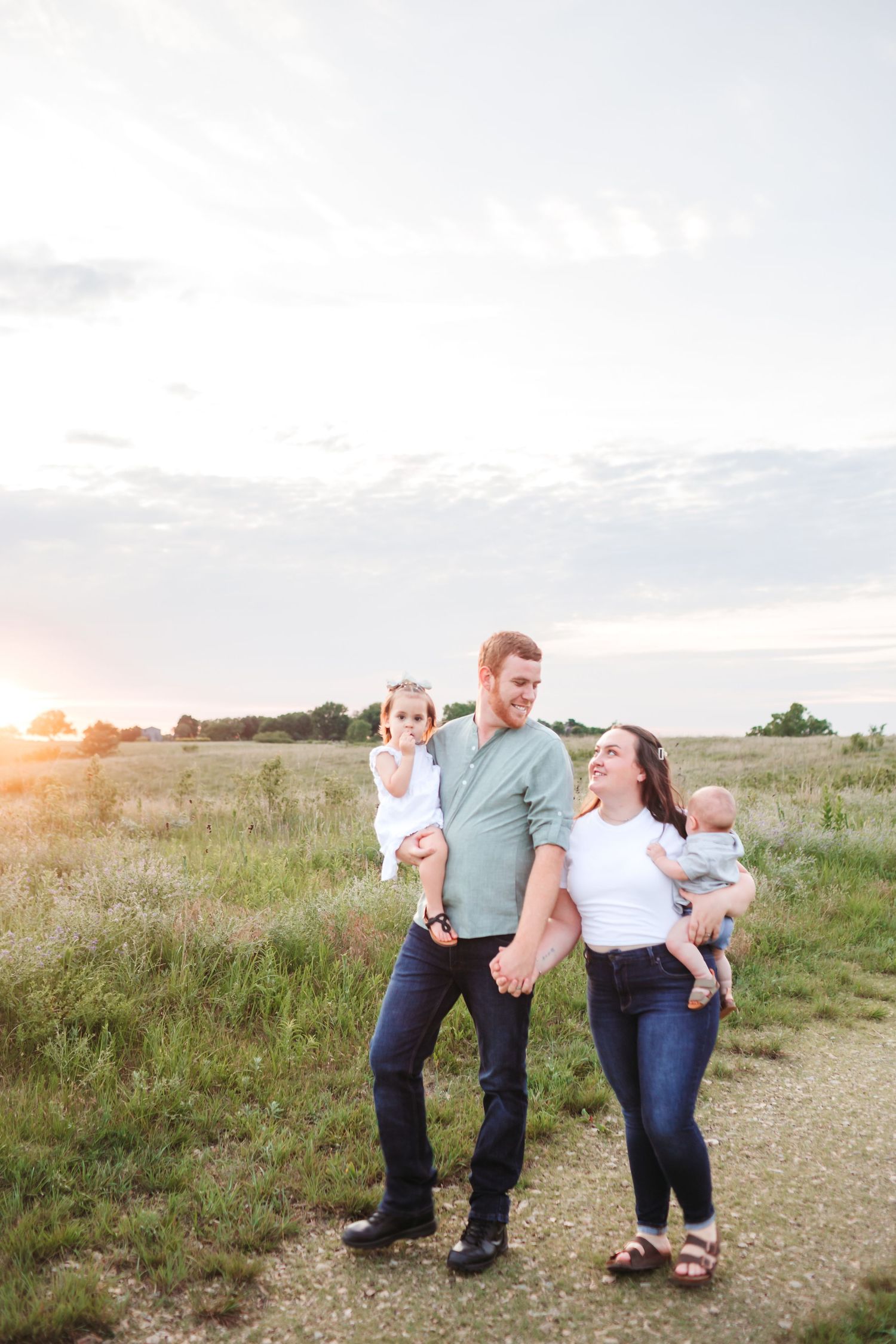 A happy family of four walks through a golden field at sunset, parents holding two young children.