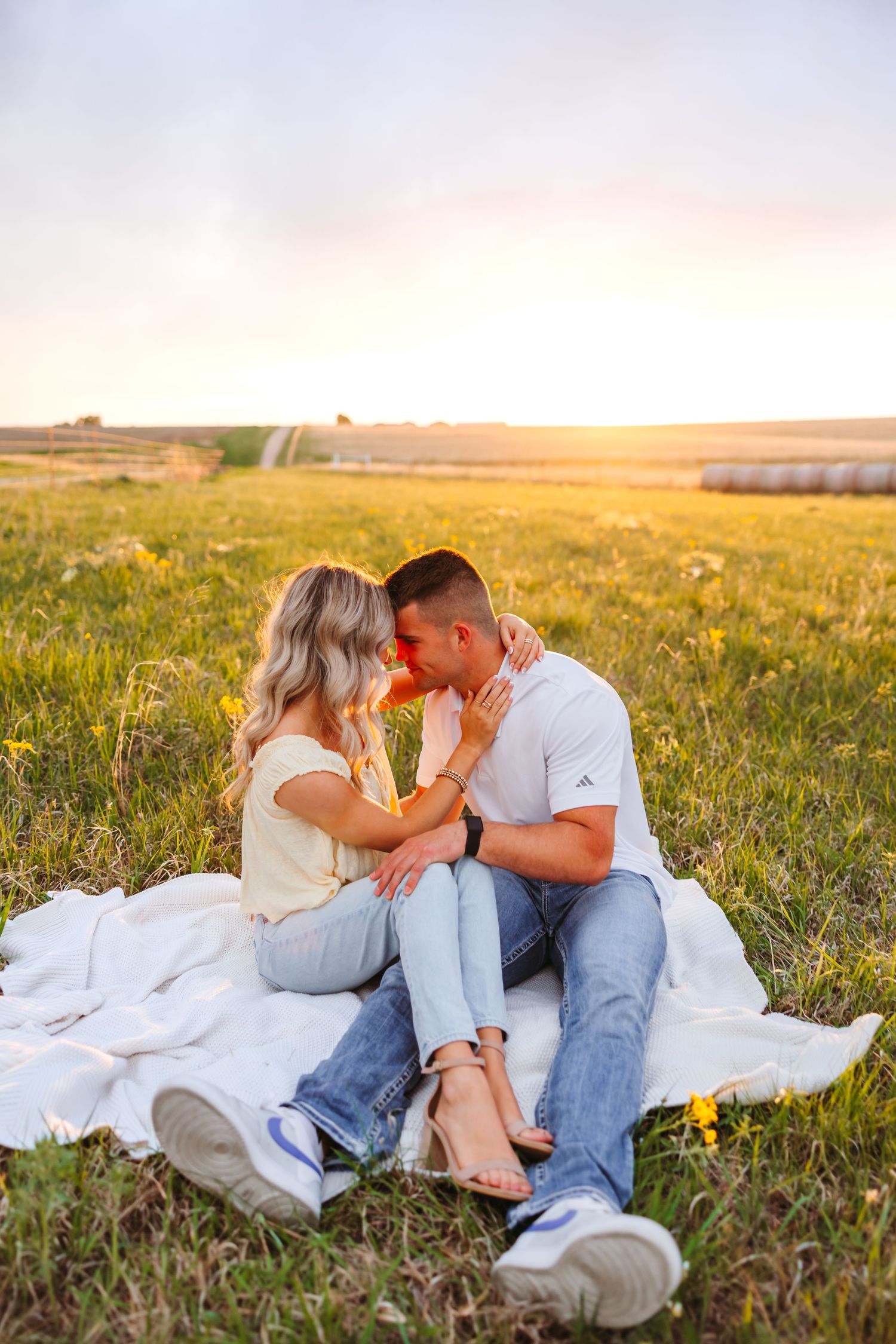 Romantic couple sitting on white blanket in golden sunset field, sharing a tender moment with hay bales in background.