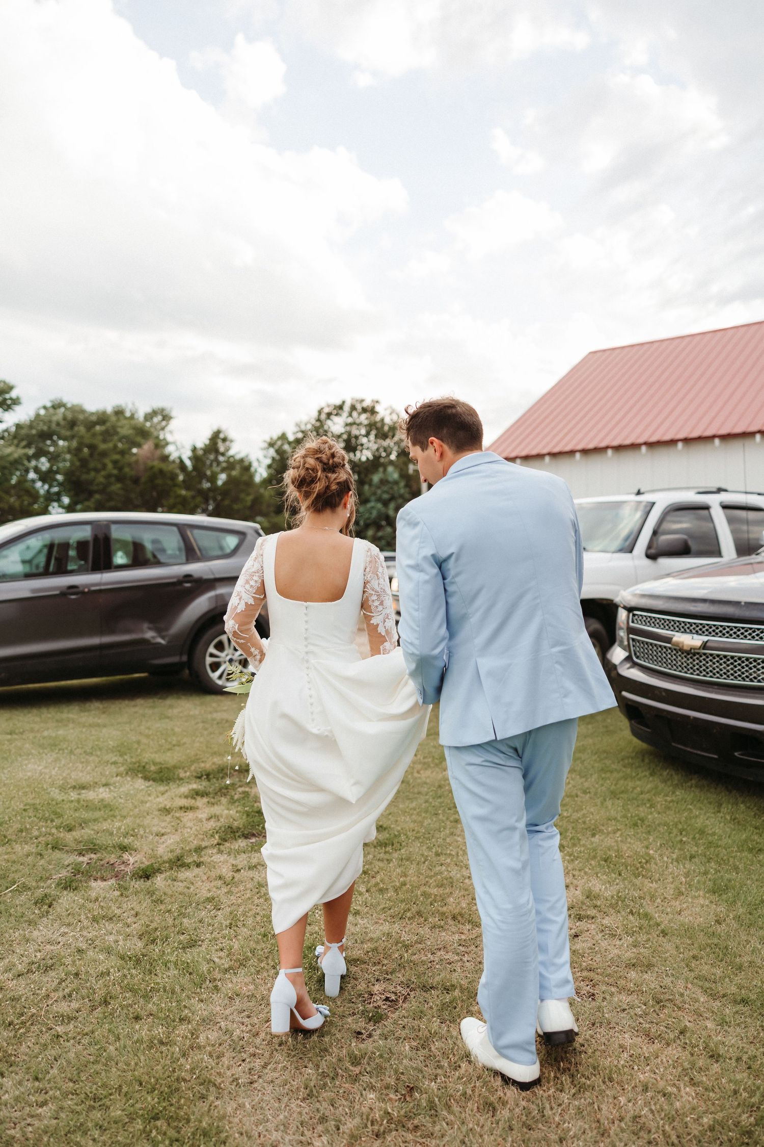 Bride in lace wedding dress and groom in light blue suit walk hand-in-hand across green lawn near parked vehicles.