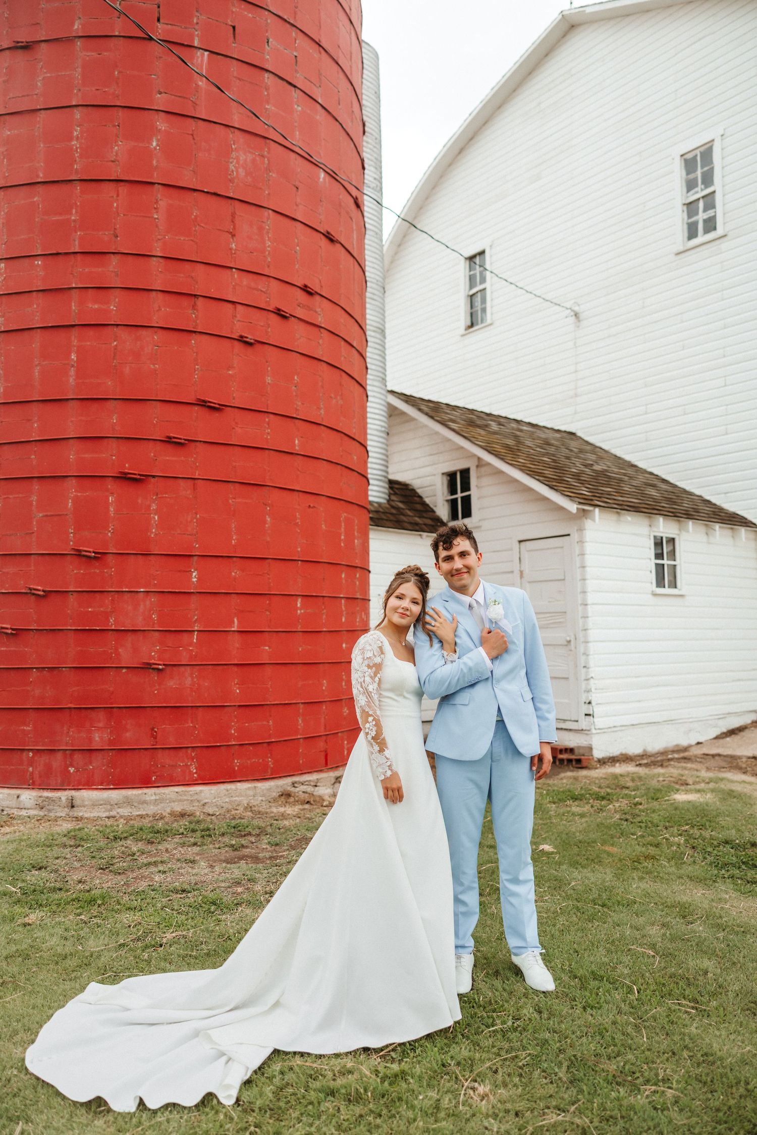 Bride and groom pose by a red silo and white barn on their rustic farm wedding day.