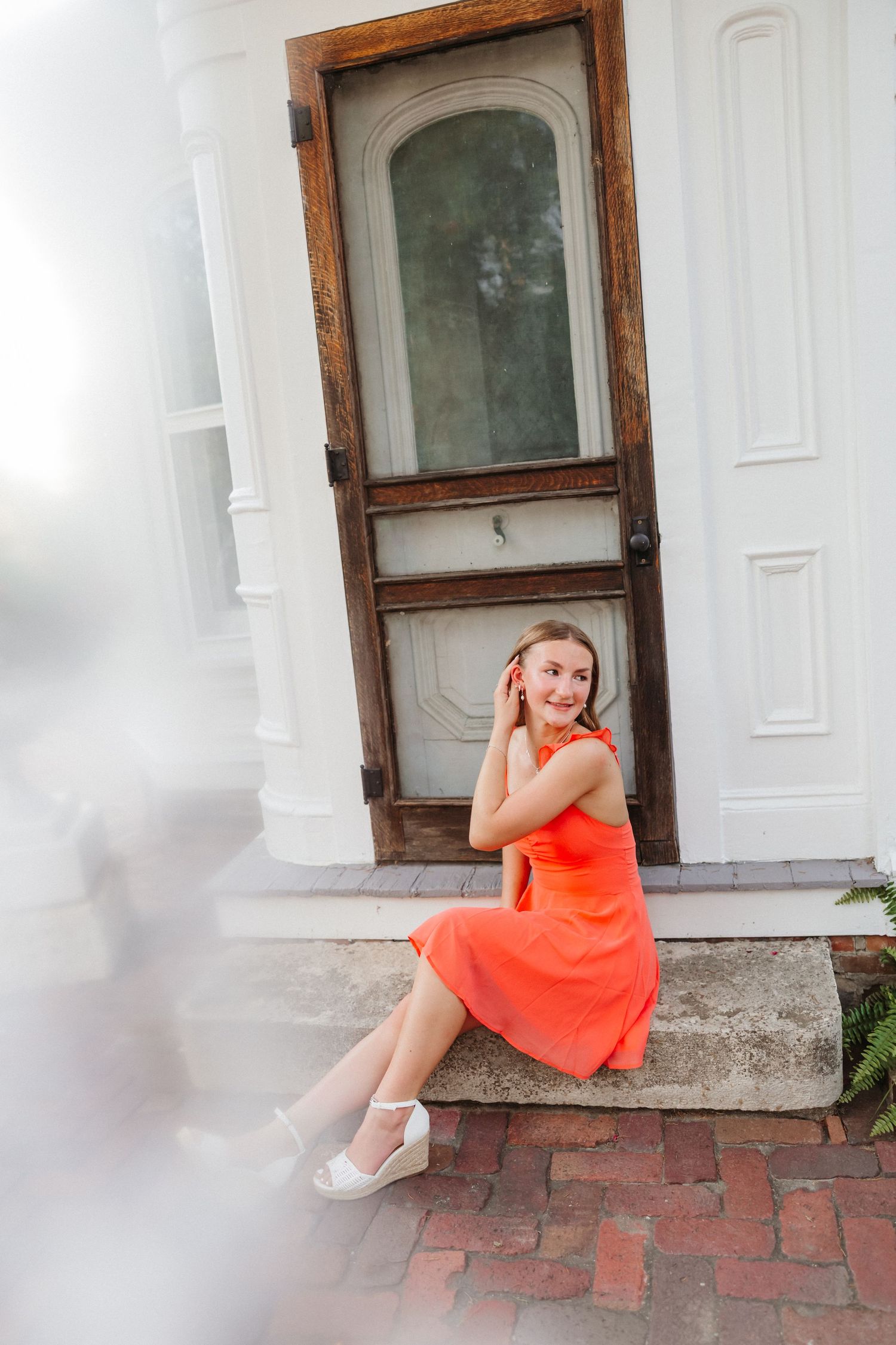 Young woman in orange dress sitting on brick steps in front of a rustic white door, smiling cheerfully.