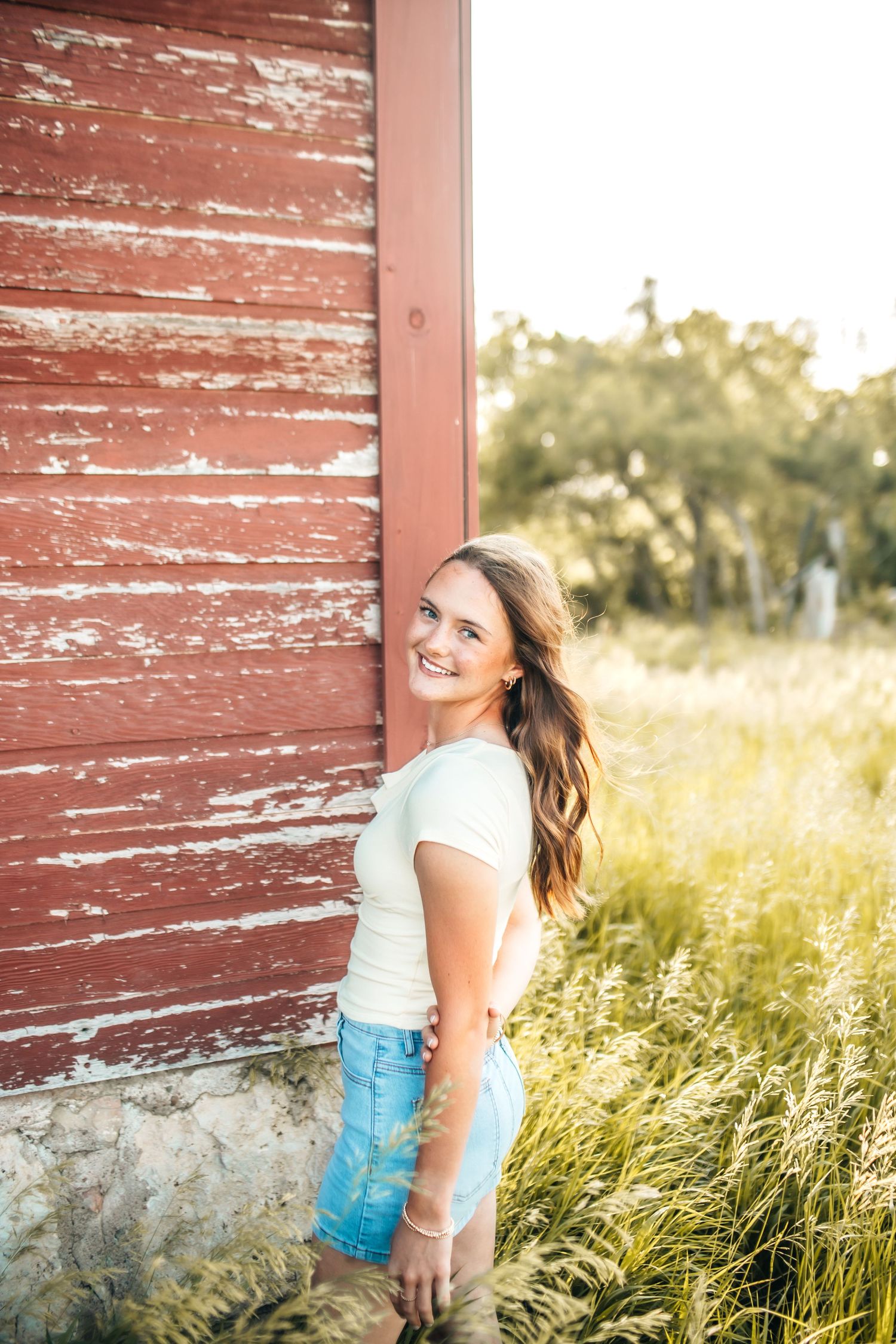 Woman in yellow shirt poses against weathered red barn wall at sunset.