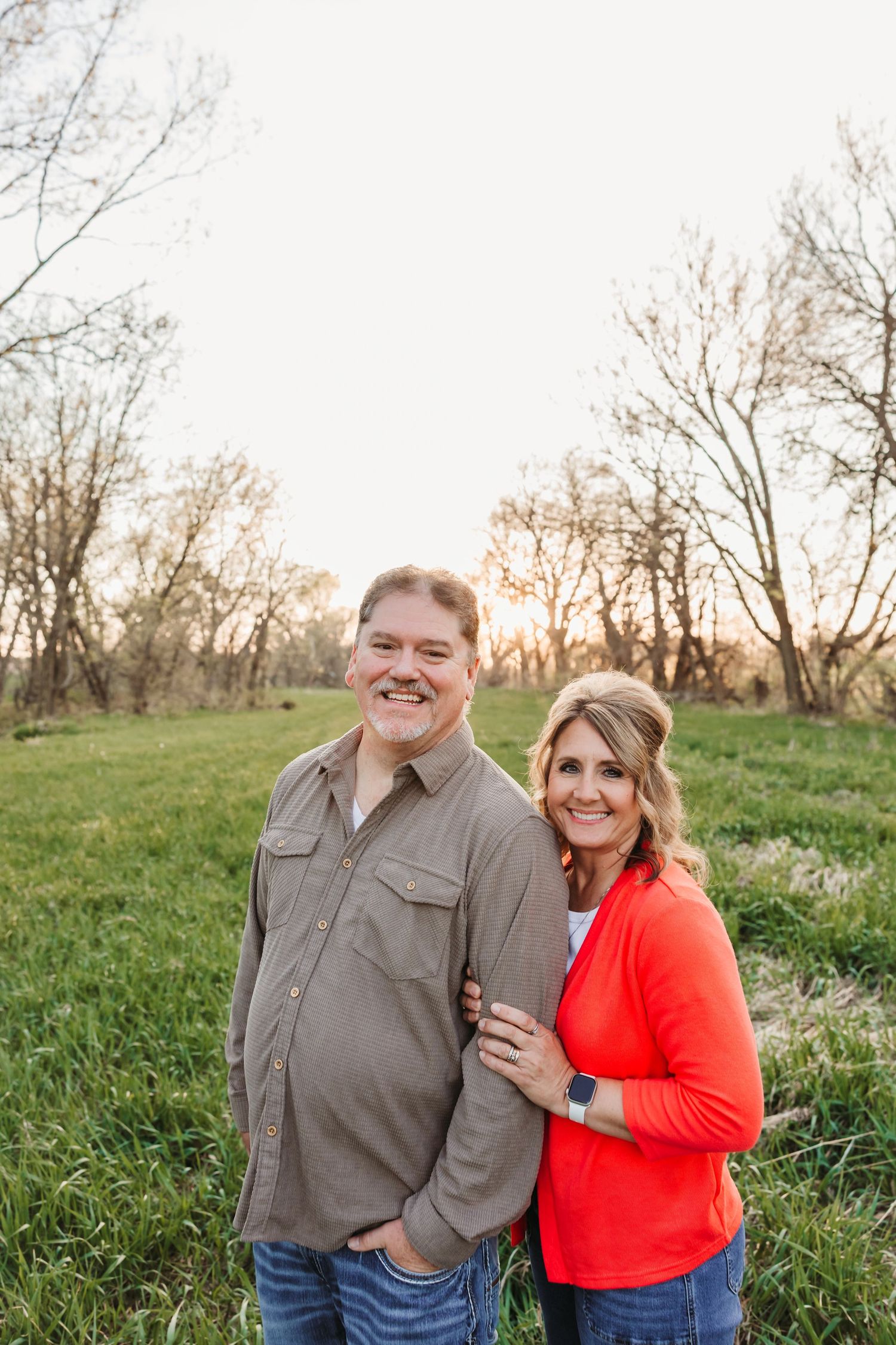 Smiling couple posing together in a green field with bare trees at sunset, man in gray shirt and woman in red cardigan.