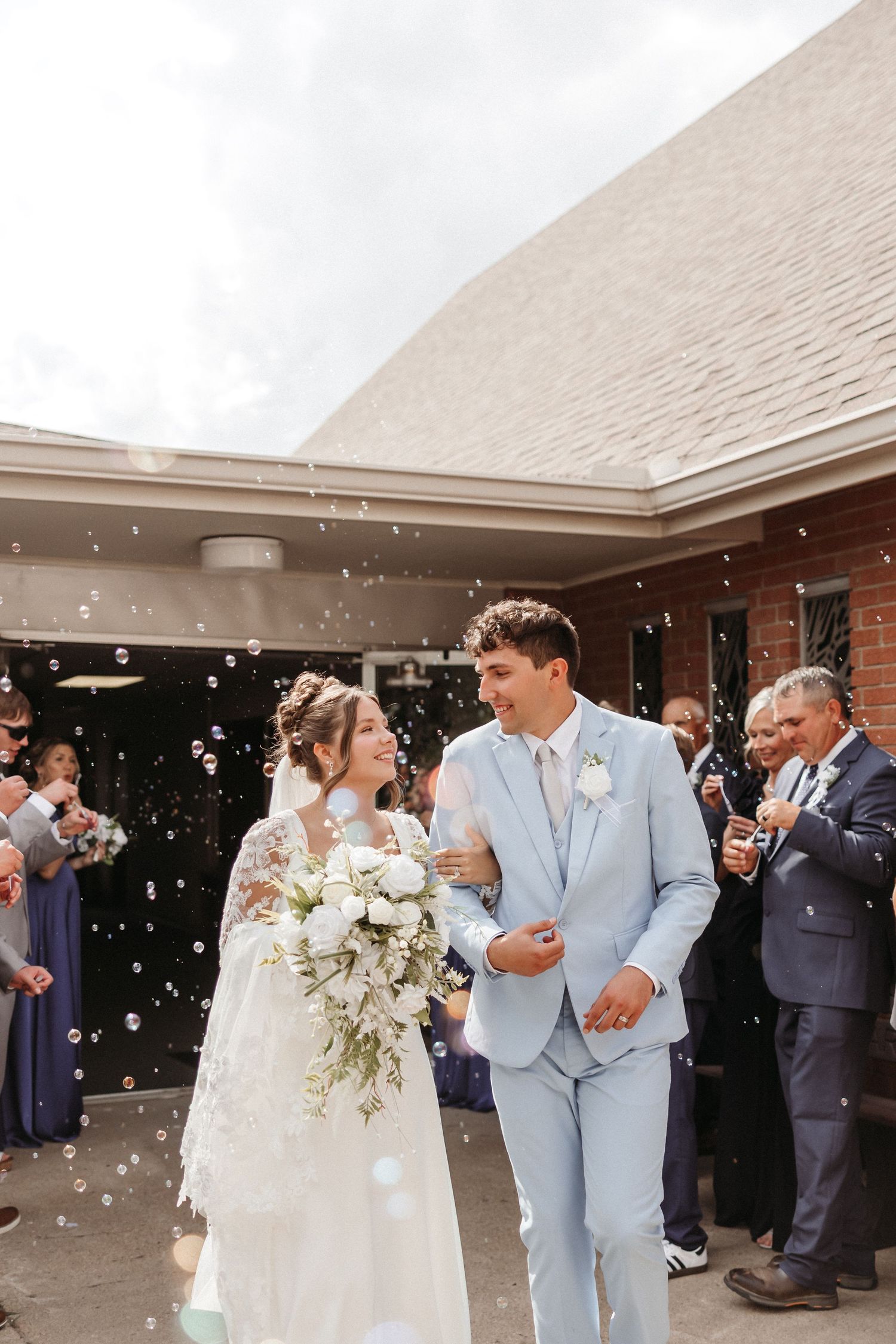 Newlywed couple exits church ceremony surrounded by bubbles, bride holding white bouquet in lace gown, groom in light blue suit.