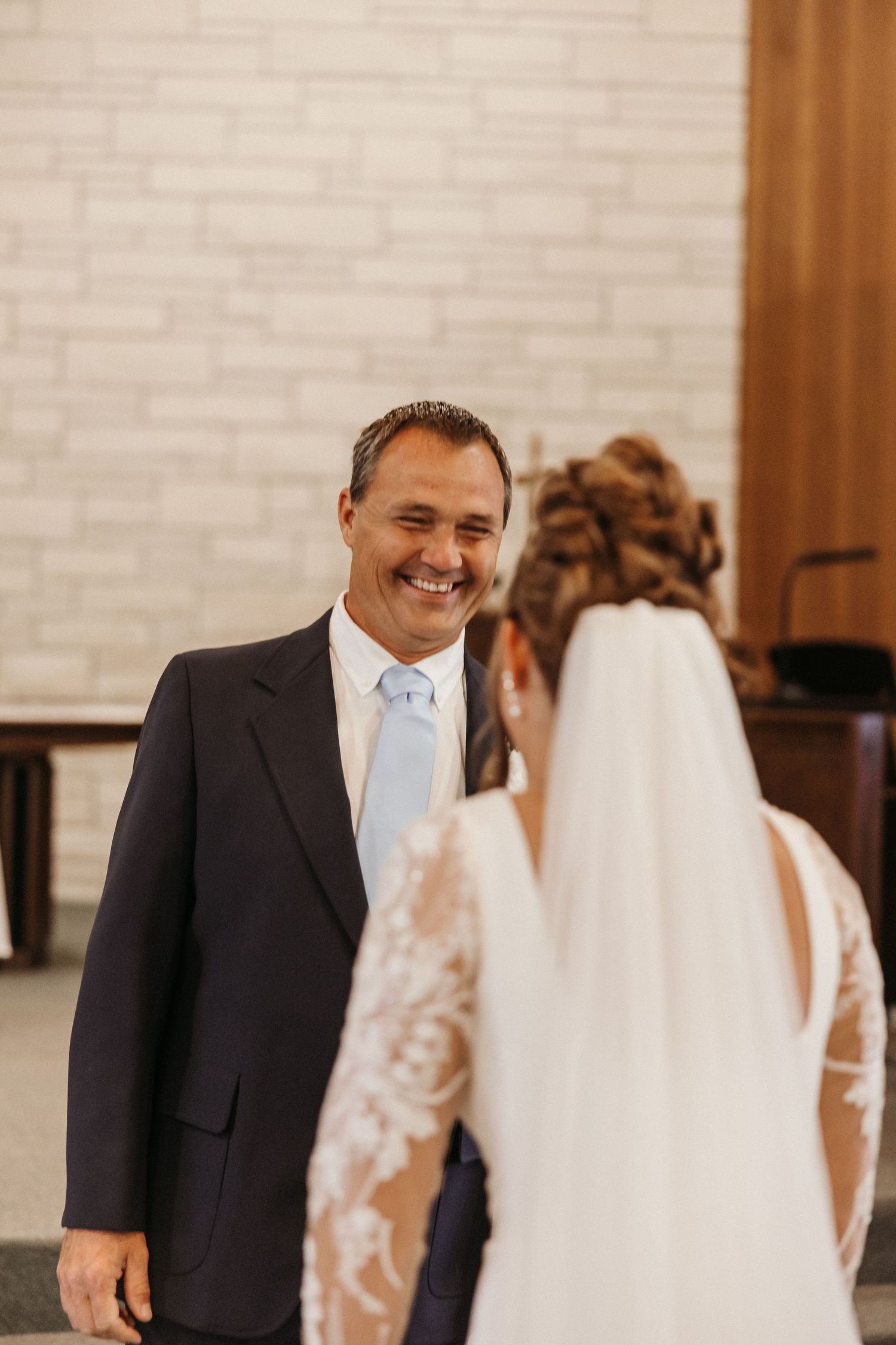 Groom smiling joyfully at bride during wedding ceremony in elegant church with white brick wall.