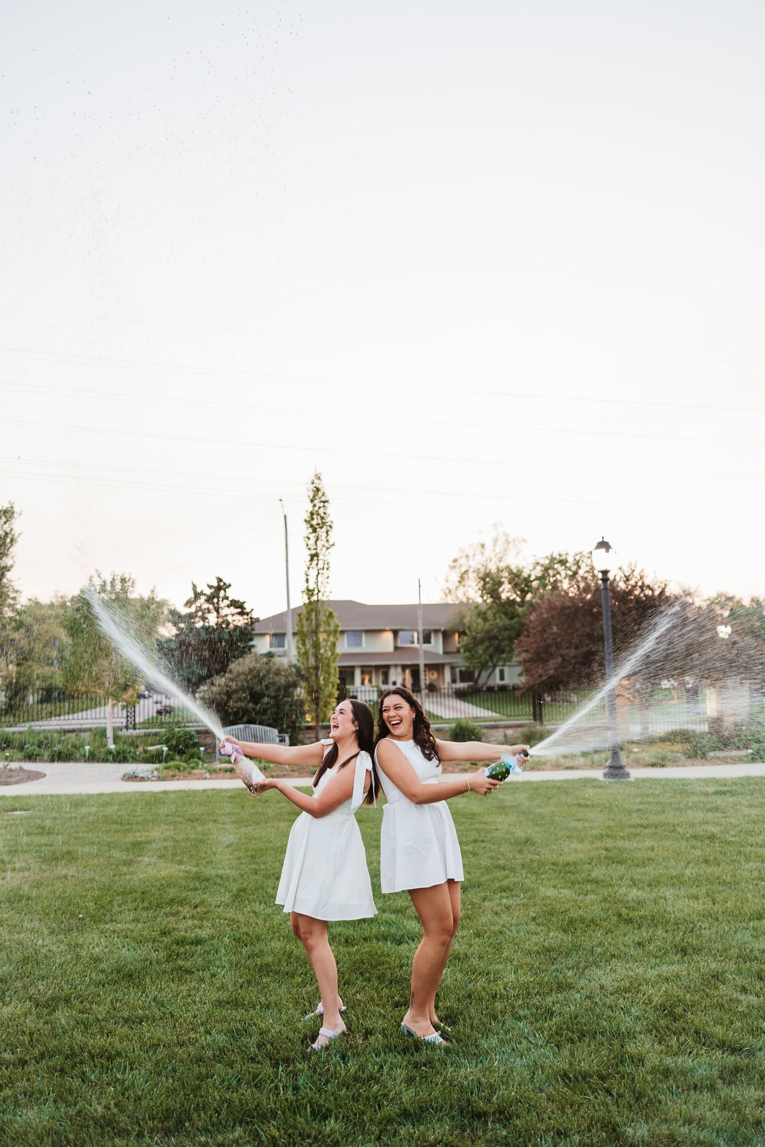 Two women in white dresses joyfully spraying champagne outdoors on green grass at dusk.