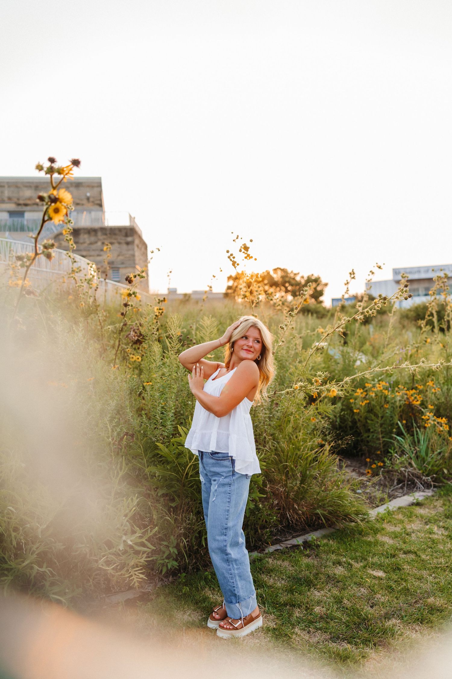 Smiling blonde woman in white top and jeans posing in a sunny garden with wildflowers at golden hour.