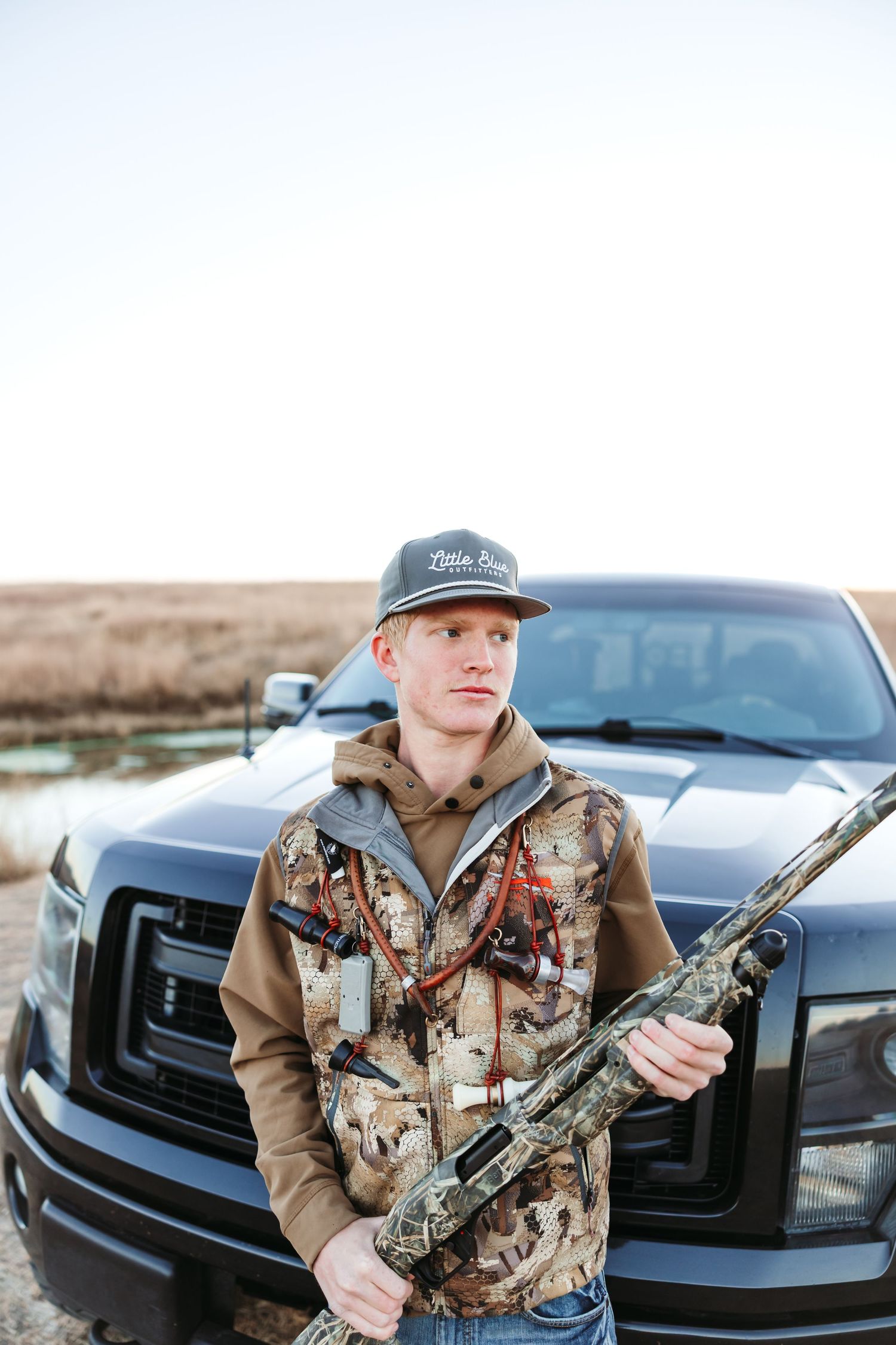 Woman in hunting gear with camouflage jacket poses with rifle near black truck.