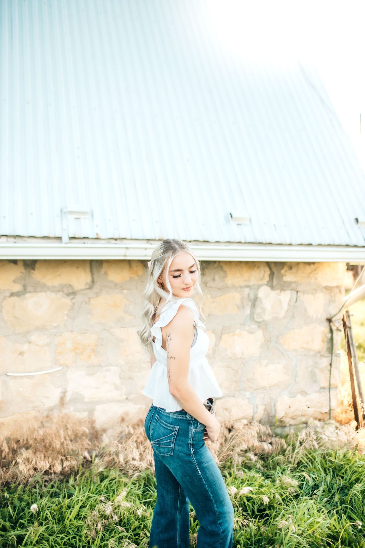 Person in white top and jeans poses near stone wall in evening light.