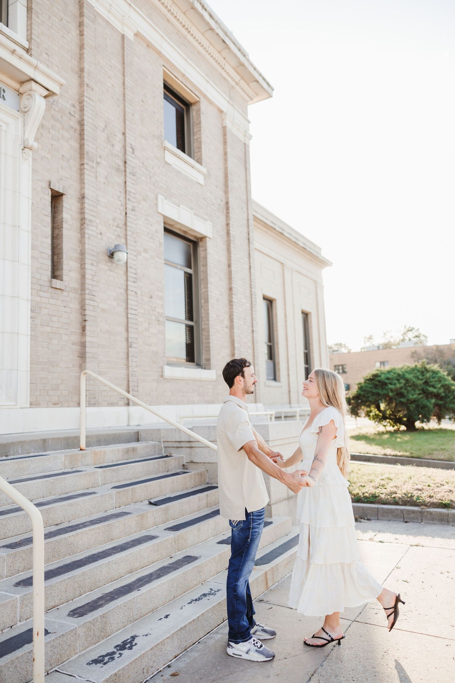 A couple holds hands while standing on the steps of a beige brick building.