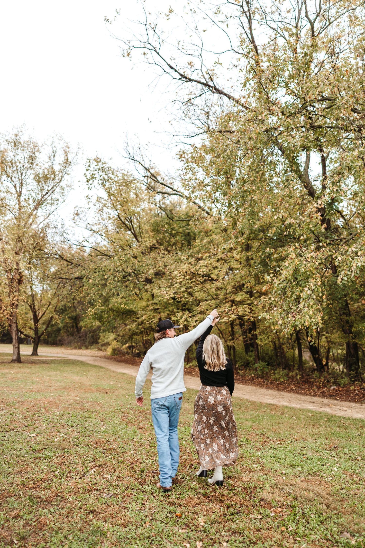 A couple walks hand in hand through a park with autumn trees overhead.