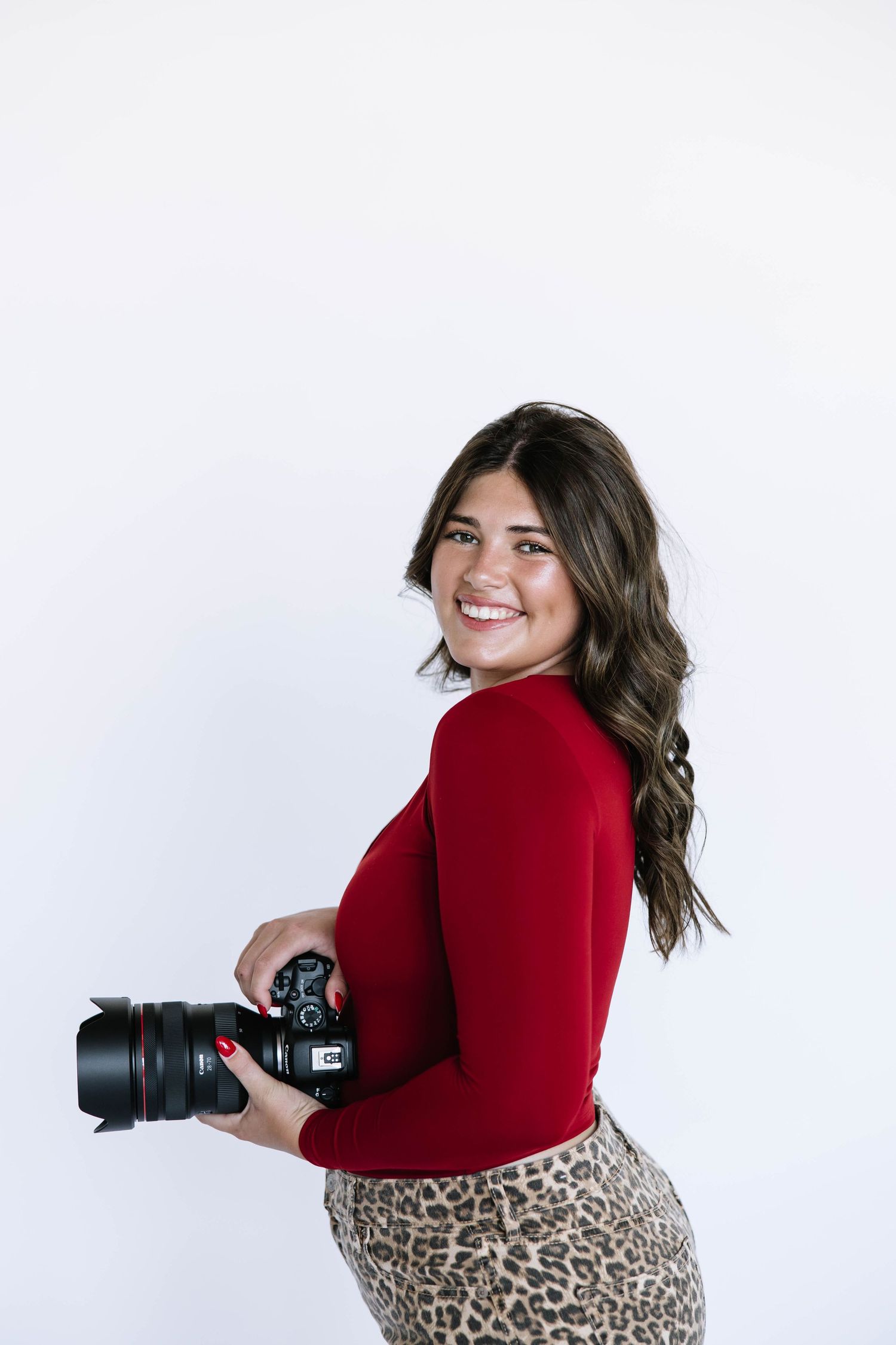 Professional photographer in red long sleeve and leopard print pants poses with camera against white backdrop.