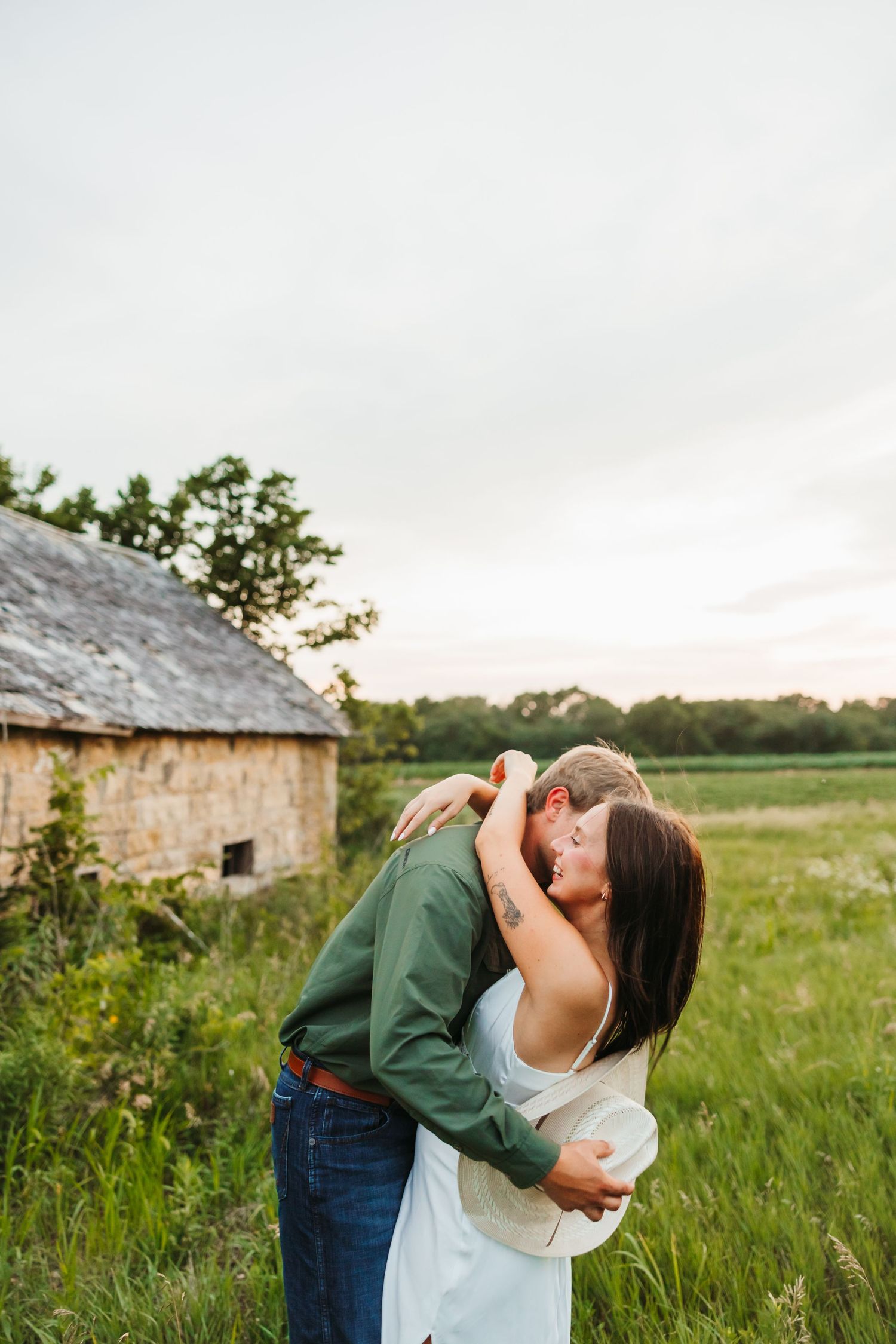 Couple sharing a romantic kiss in a green field near a rustic stone barn at sunset.