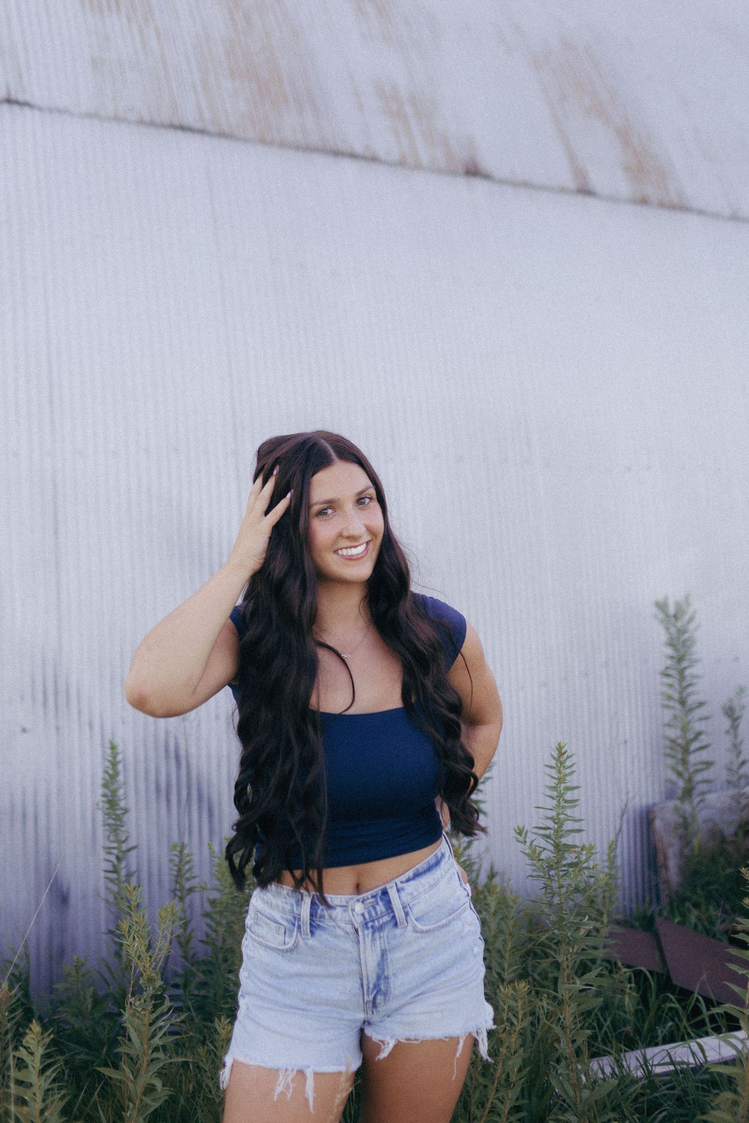 Young woman with long wavy dark hair in blue crop top and denim shorts posing outdoors near green plants and metal wall.