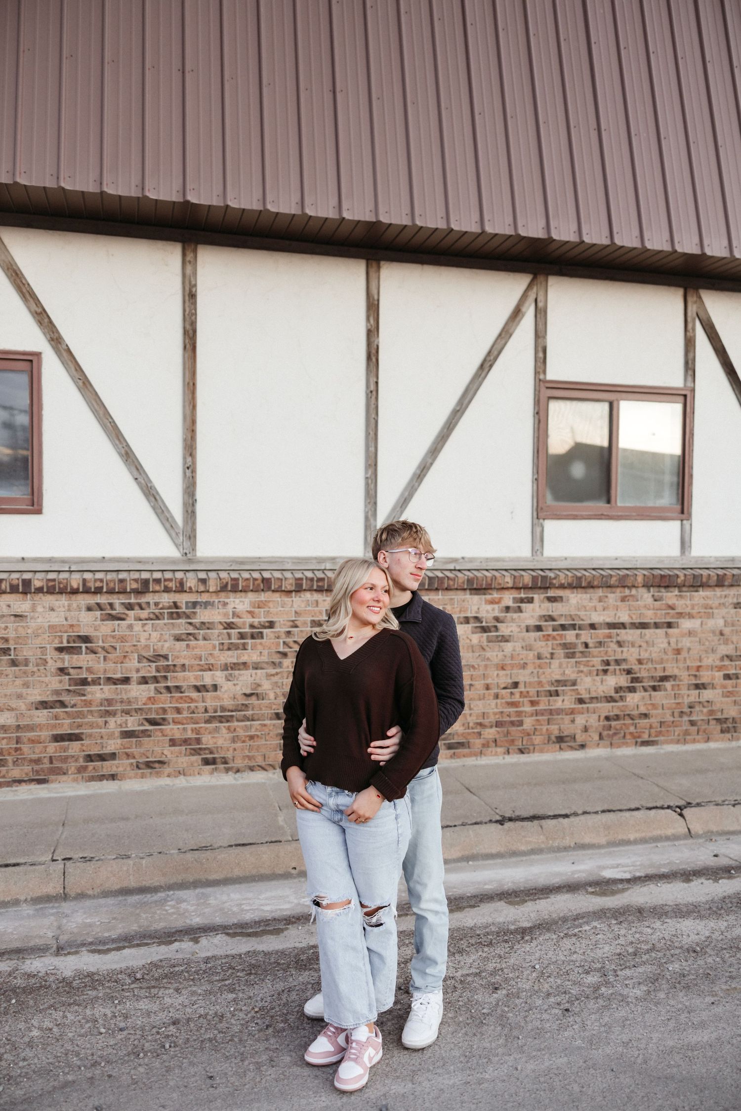 A couple embracing and smiling in front of a brick building, wearing matching dark sweaters and light jeans.