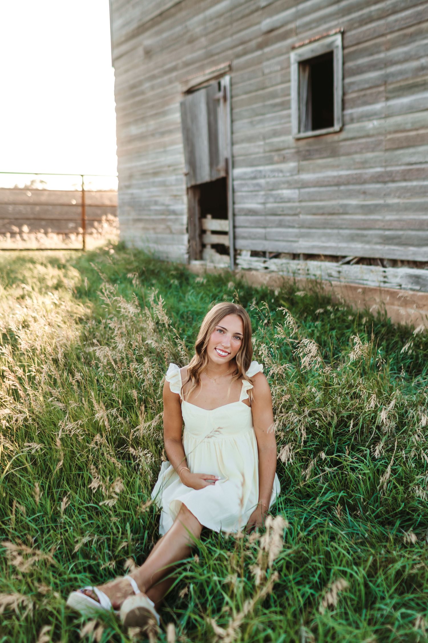 Smiling woman in white dress sitting in tall grass beside a rustic wooden barn at golden hour.