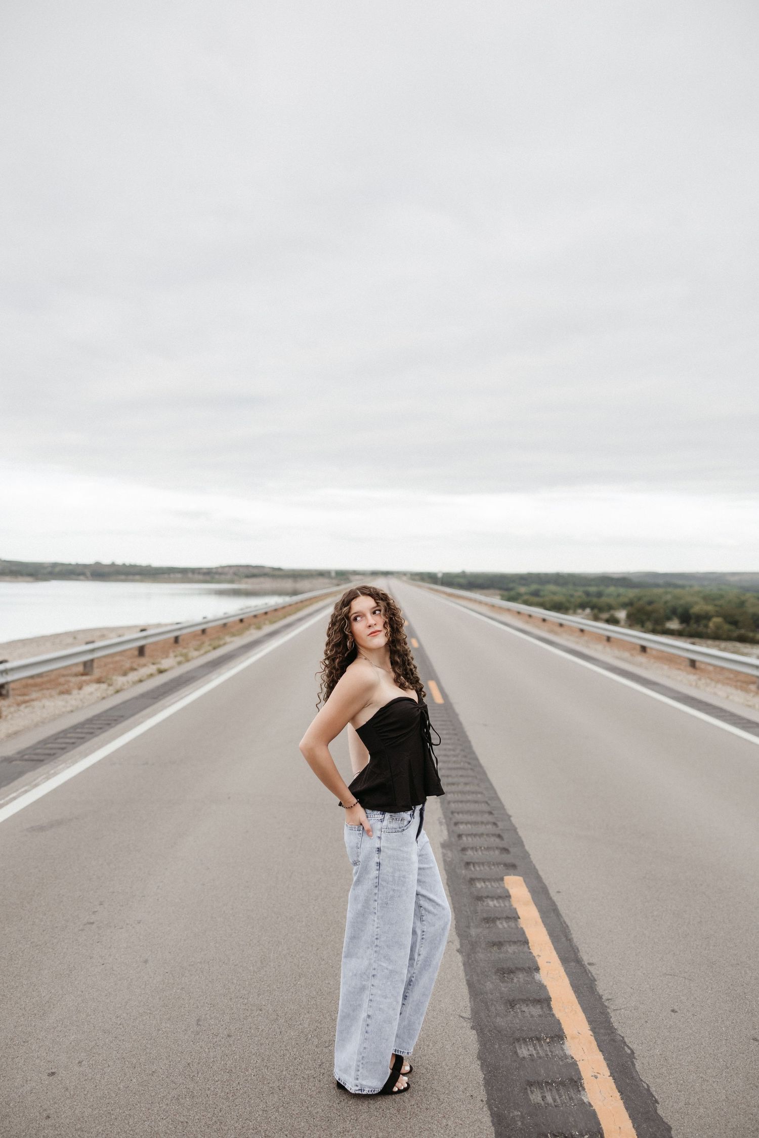 Young woman in black strapless top and wide-leg jeans posing on an empty bridge road under overcast skies.