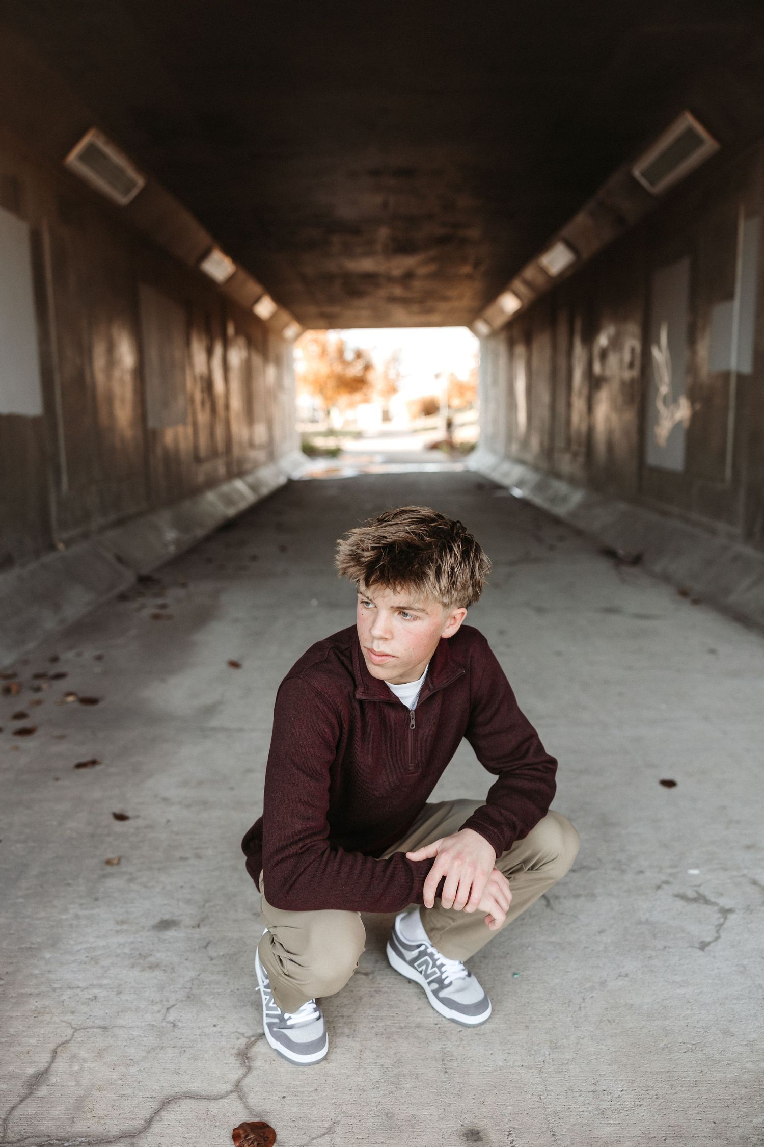 Person in burgundy sweater crouches in dimly lit concrete tunnel.