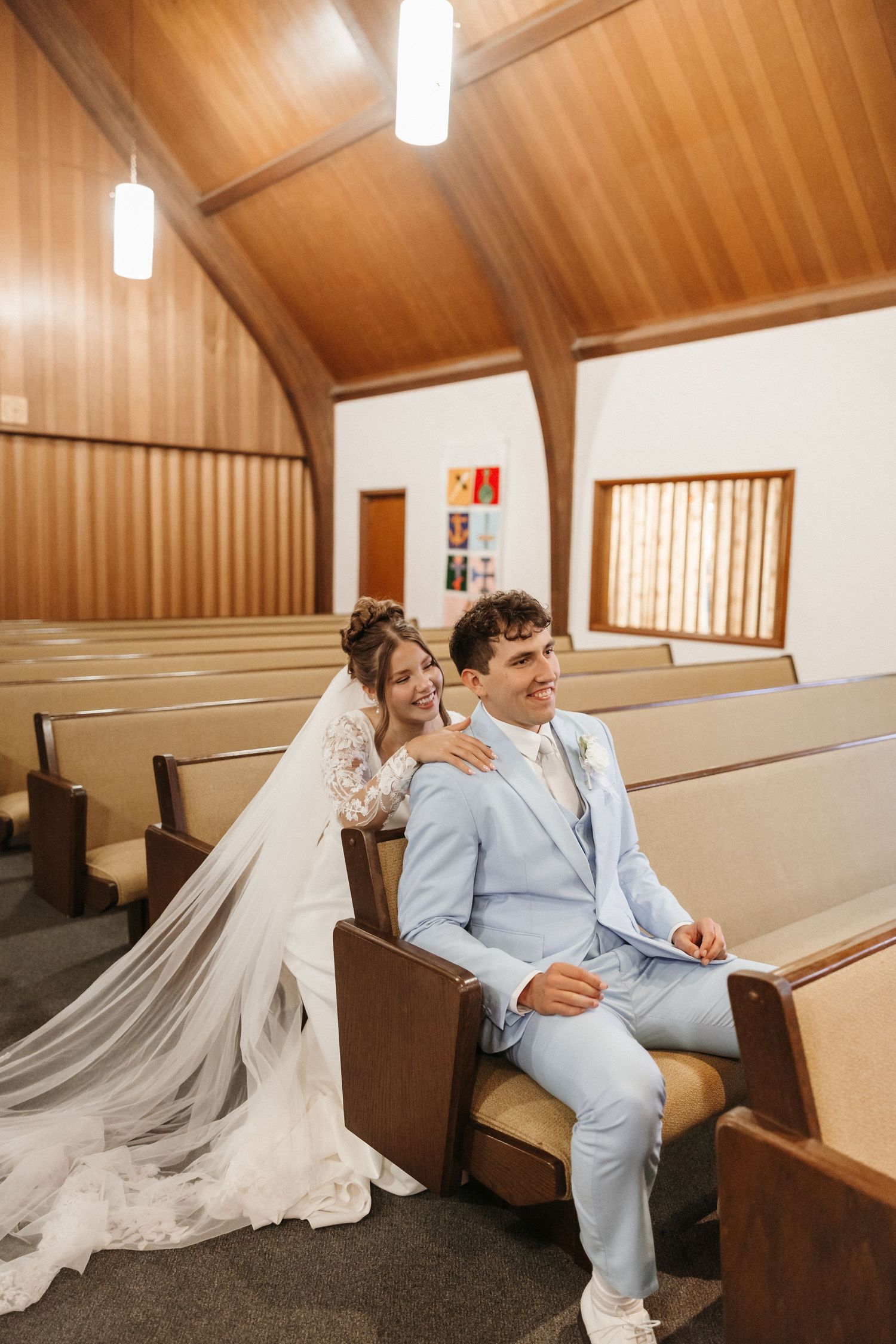 Bride in white gown and groom in light blue suit sitting together in wooden church pews on their wedding day.
