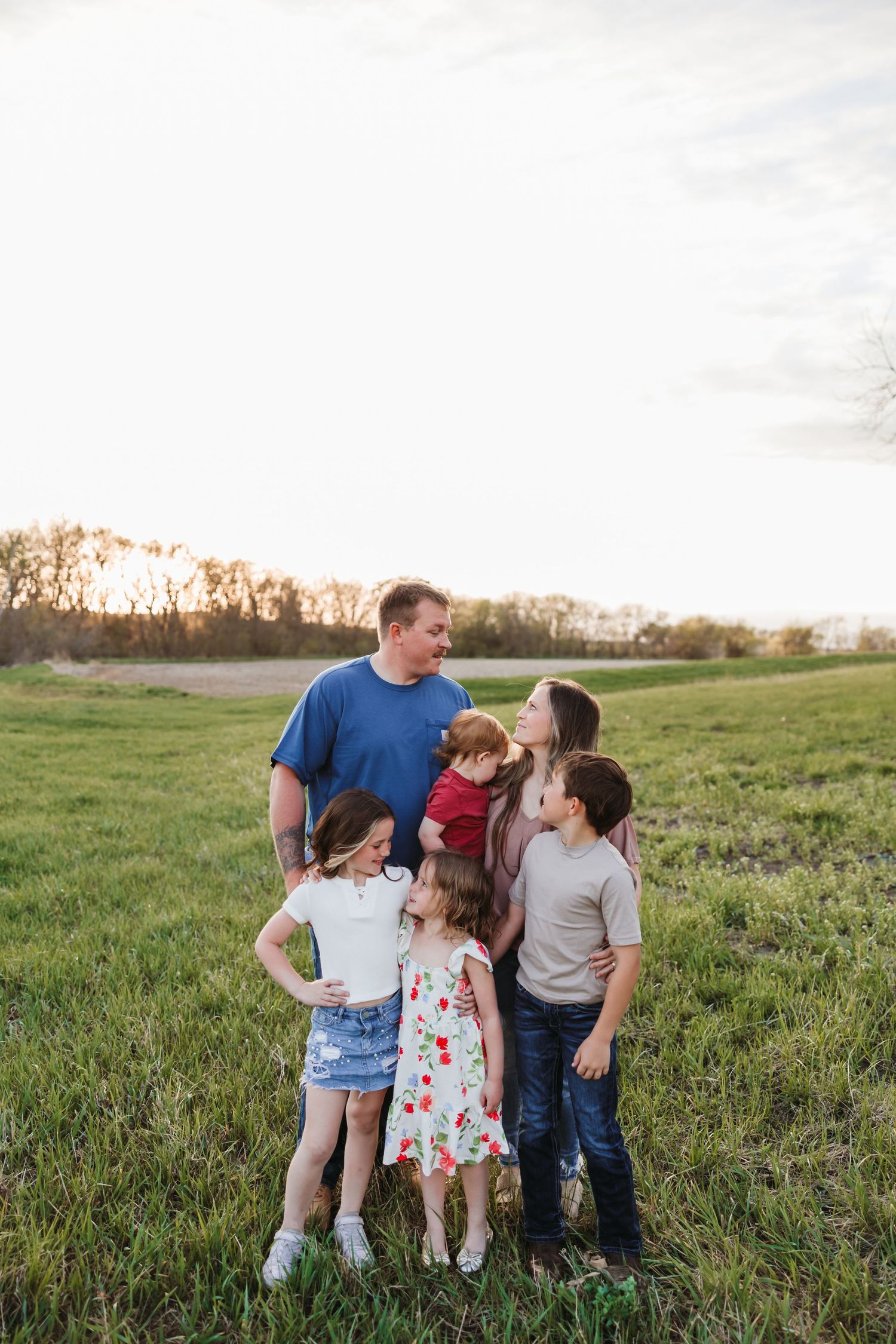 Family of six posing in a green field at golden hour, parents and four children smiling together outdoors.