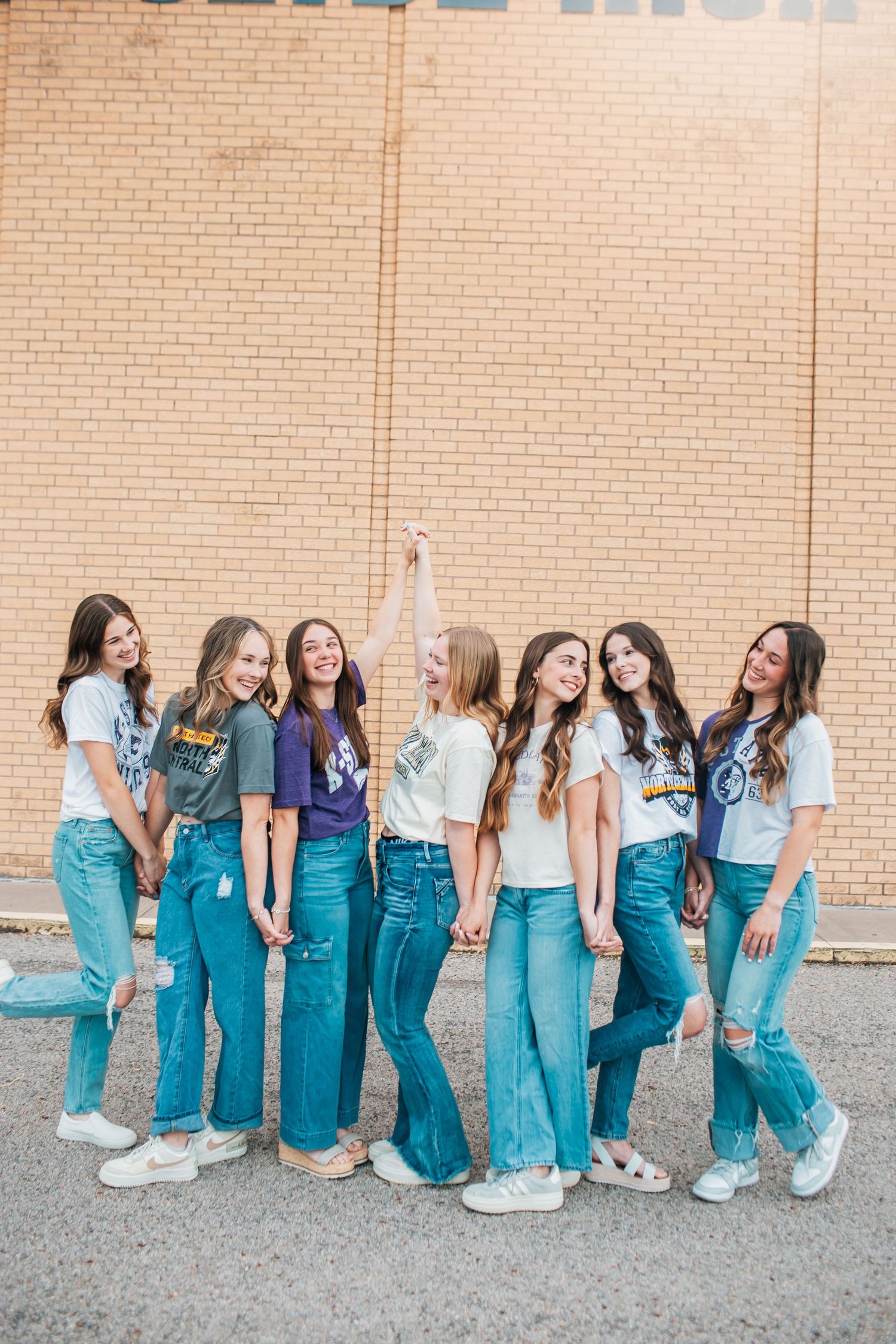 Group of senior girls in college tees and jeans posed against brick wall at high school.