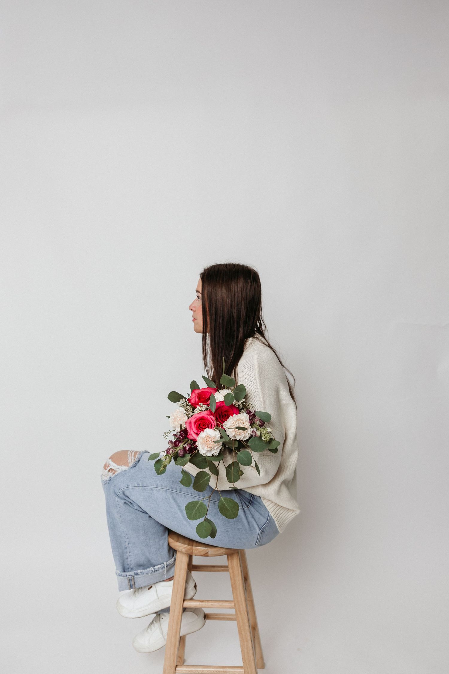 Side profile on wooden stool holding floral arrangement against white wall.