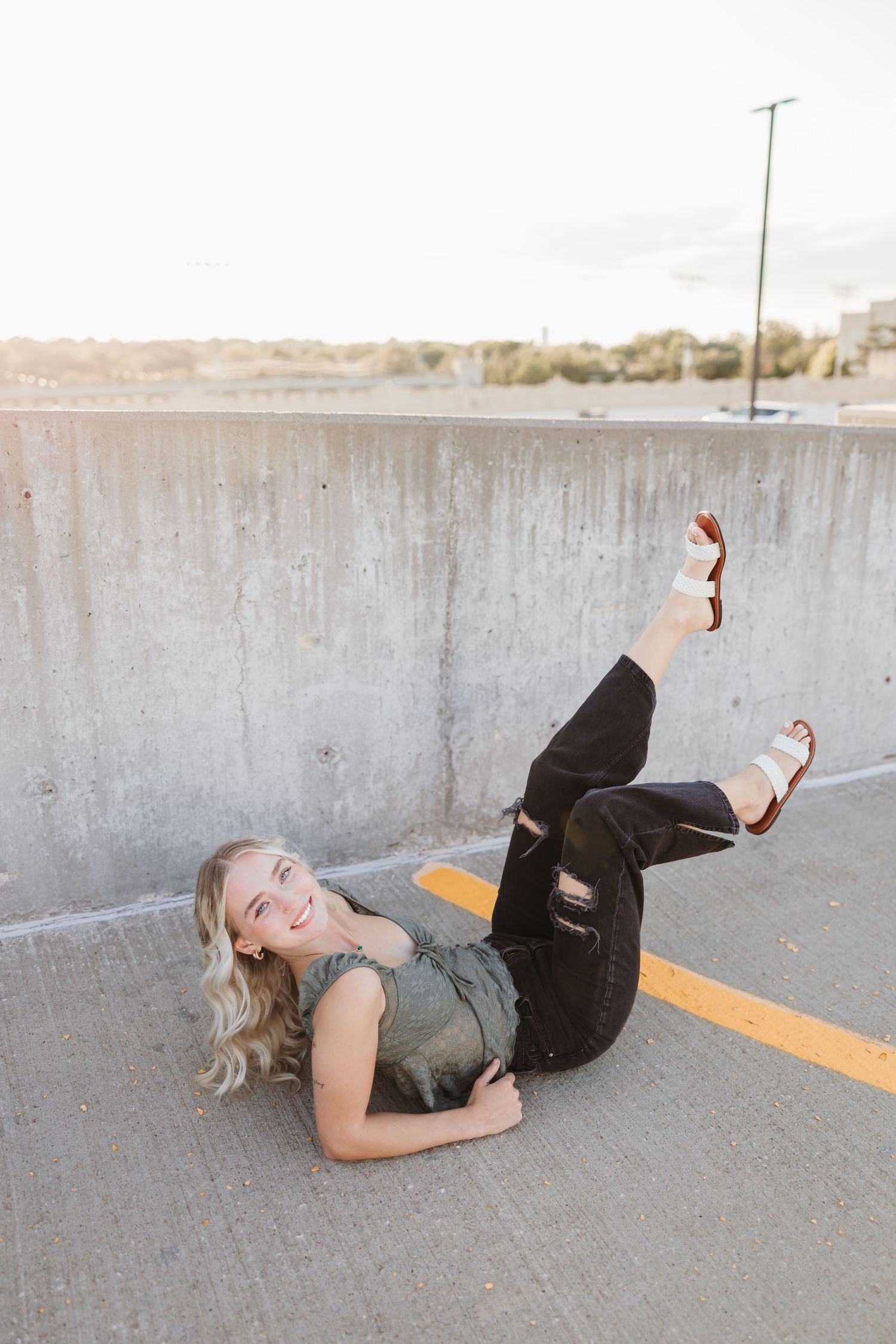 Young blonde woman posing on parking garage floor, wearing black jeans and sandals, smiling with legs raised.