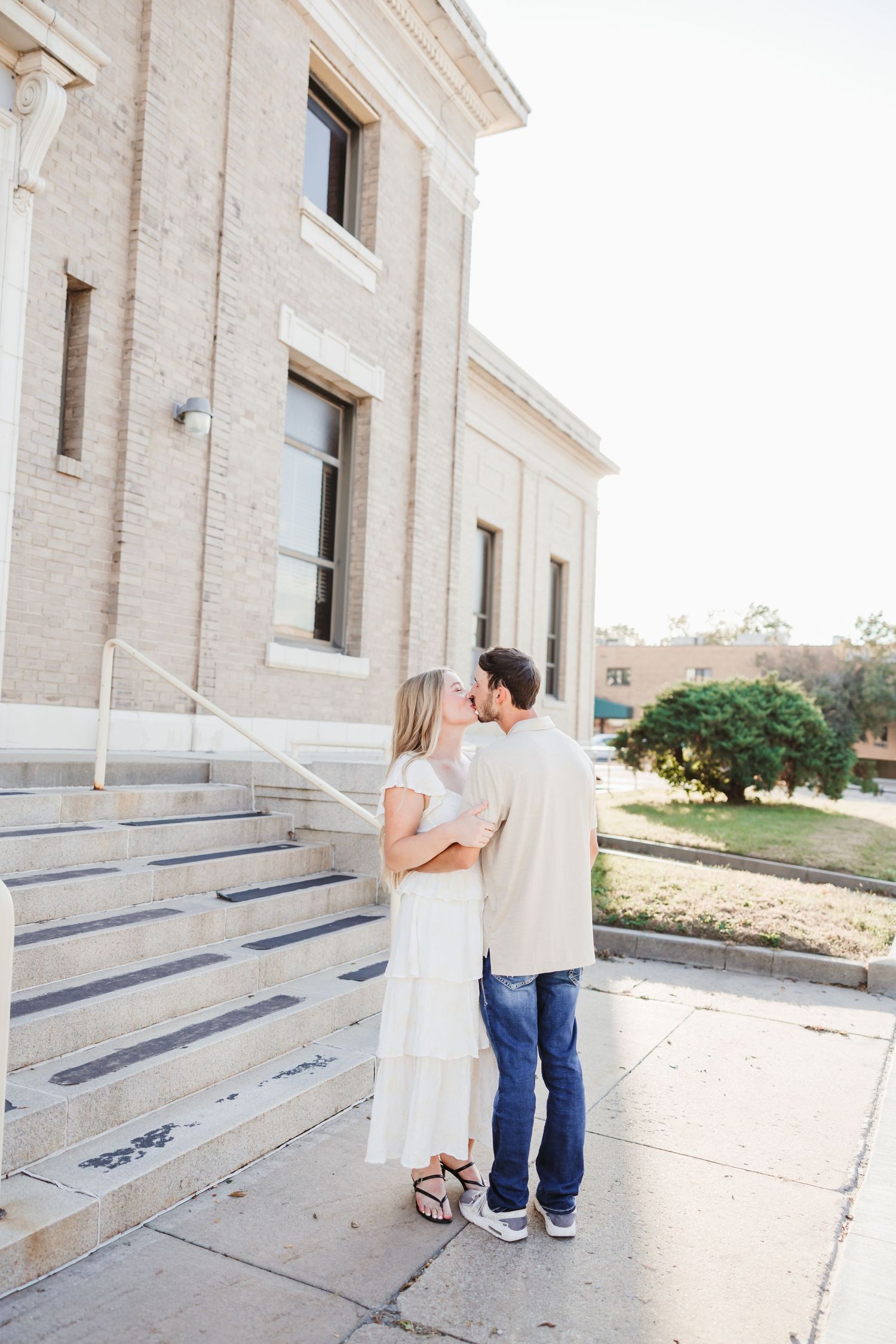Couple sharing a kiss outside a historic brick building with stone steps in warm sunlight.